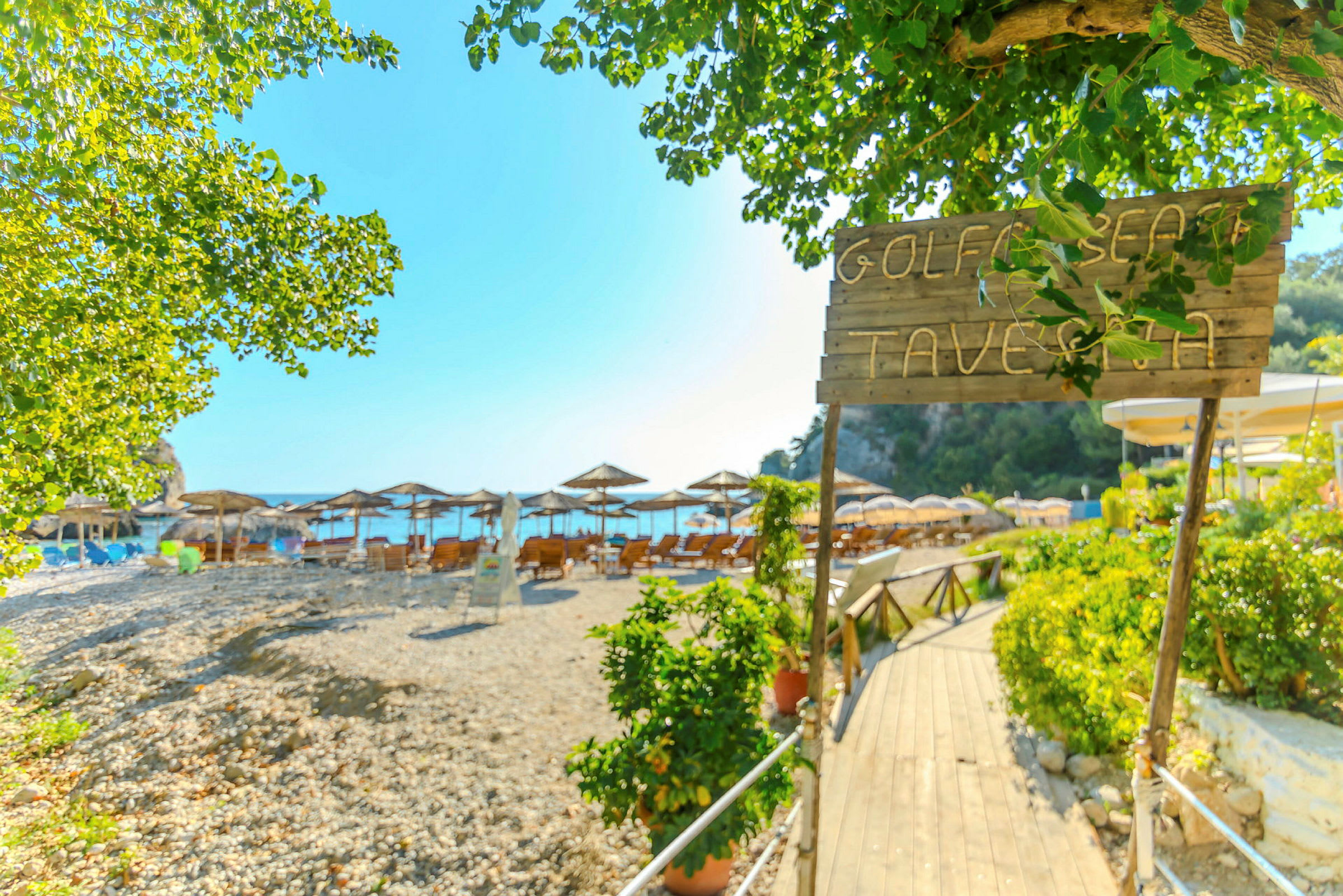 Skilt med teksten 'Golf Beach Taverna' ved indgangen til en strand med parasoller og liggestole. (AI-genereret tekst)