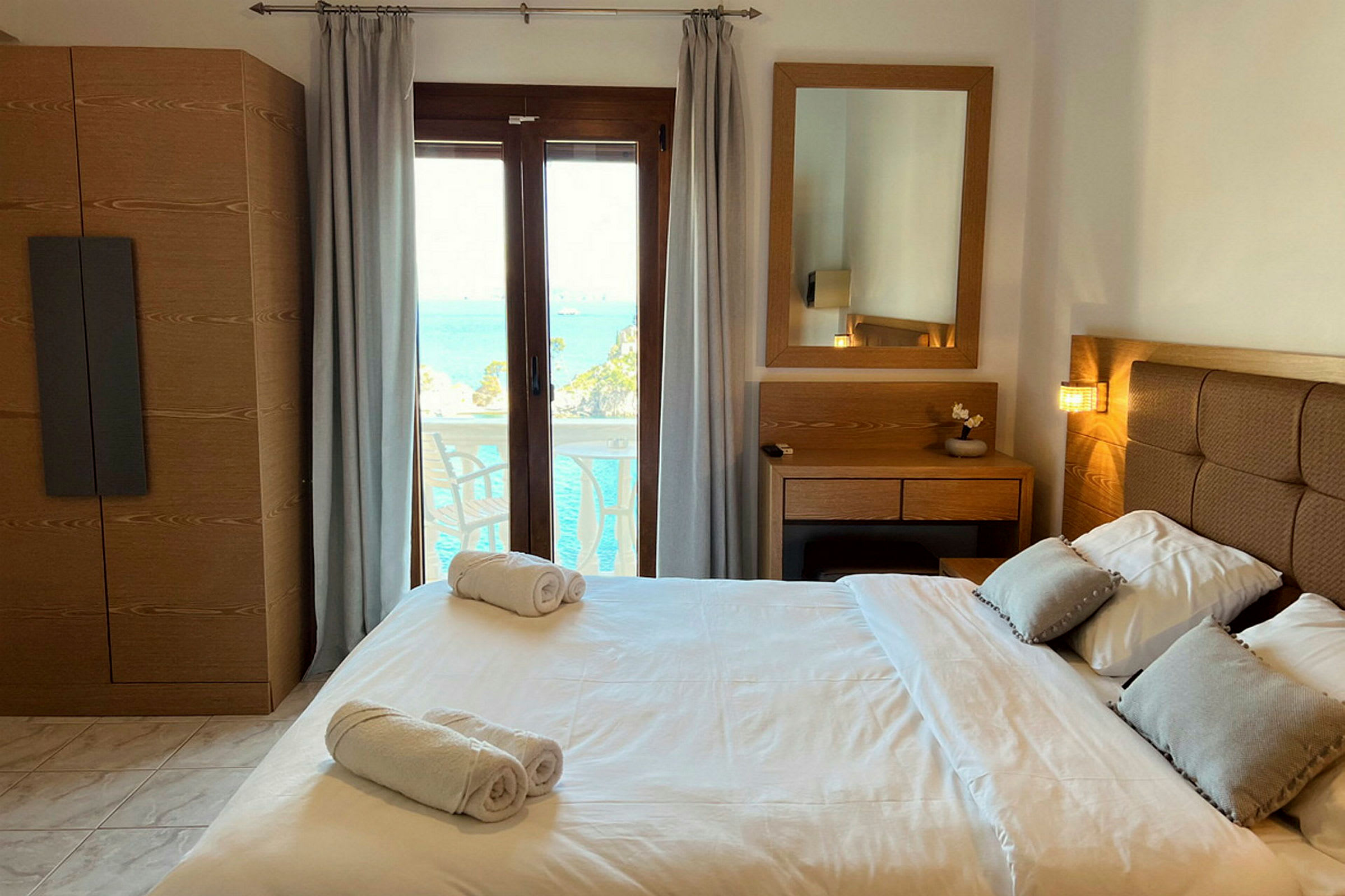 Et hotelværelse med en dobbeltseng, natbord, spejl og udsigt over havet gennem en glasdør. (AI-genereret tekst)