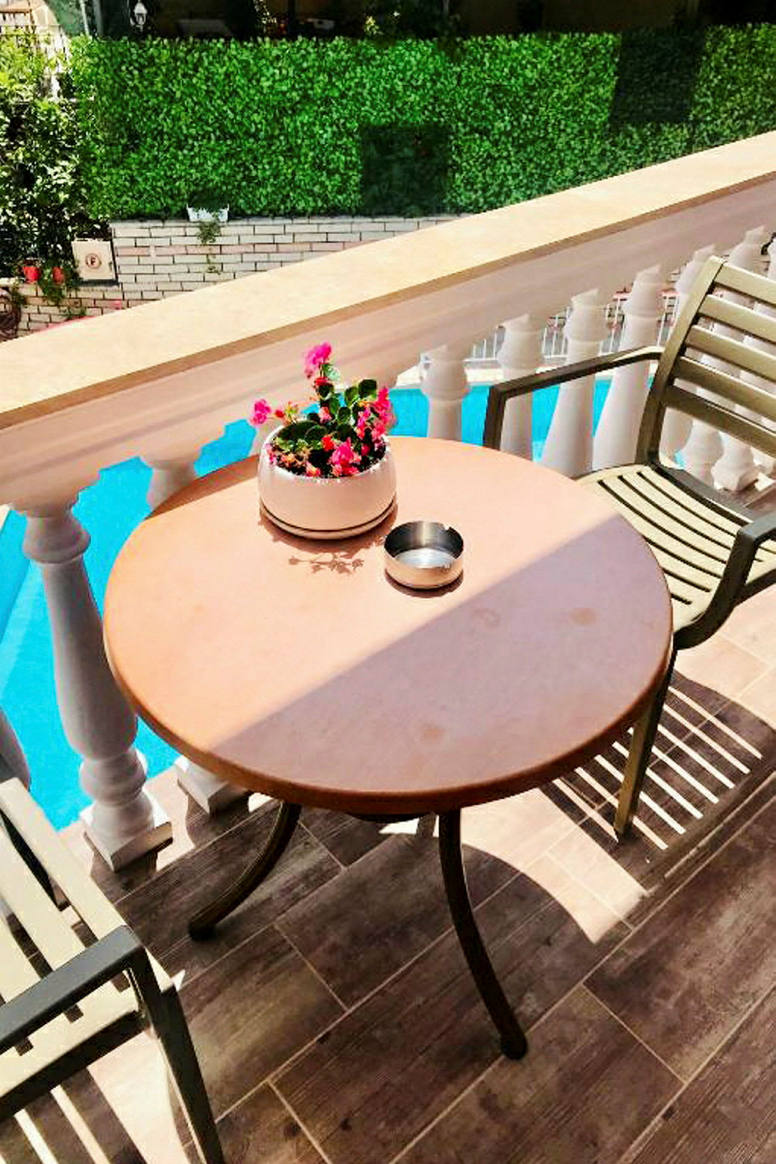 En terrasse med et rundt bord, en stol og en blomsterkrukke. I baggrunden ses et gelænder og en pool. (AI-genereret tekst)