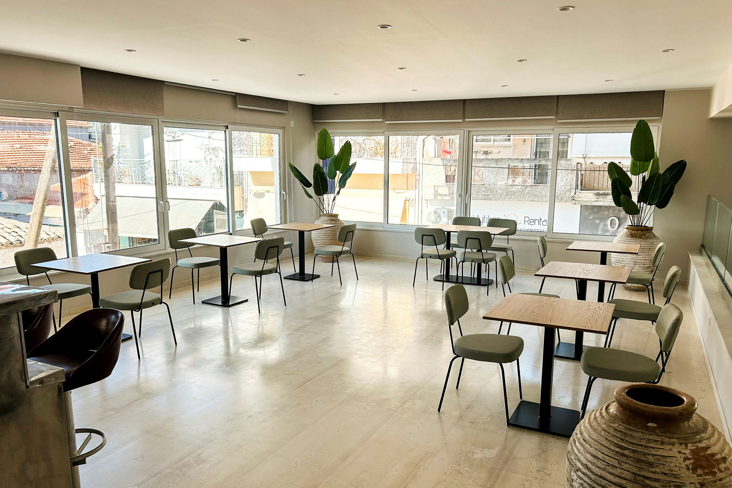 En lys café med borde og stole samt store vinduer, der lukker naturligt lys ind. Flere potteplanter pynter rummet. (AI-genereret tekst)