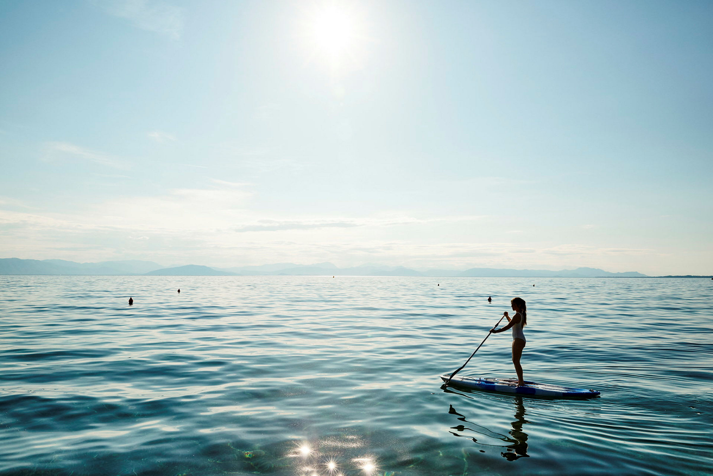 Kvinde, der står på et paddleboard på en rolig sø med solskin og bjerge i baggrunden. (AI-genereret tekst)
