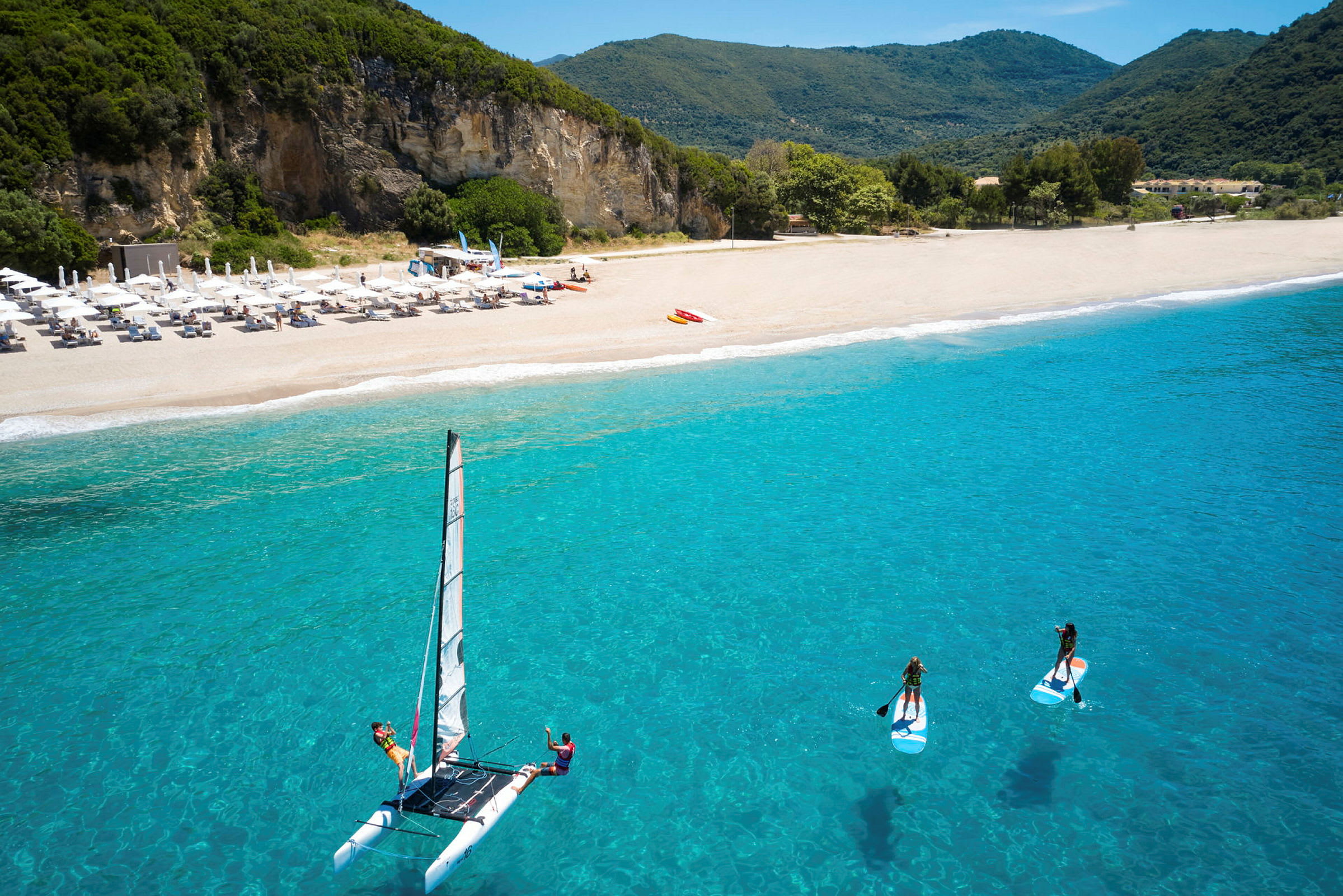 En strand med parasoller, klipper i baggrunden og aktiviteter på vandet, inklusive sejlads og paddleboarding. (AI-genereret tekst)