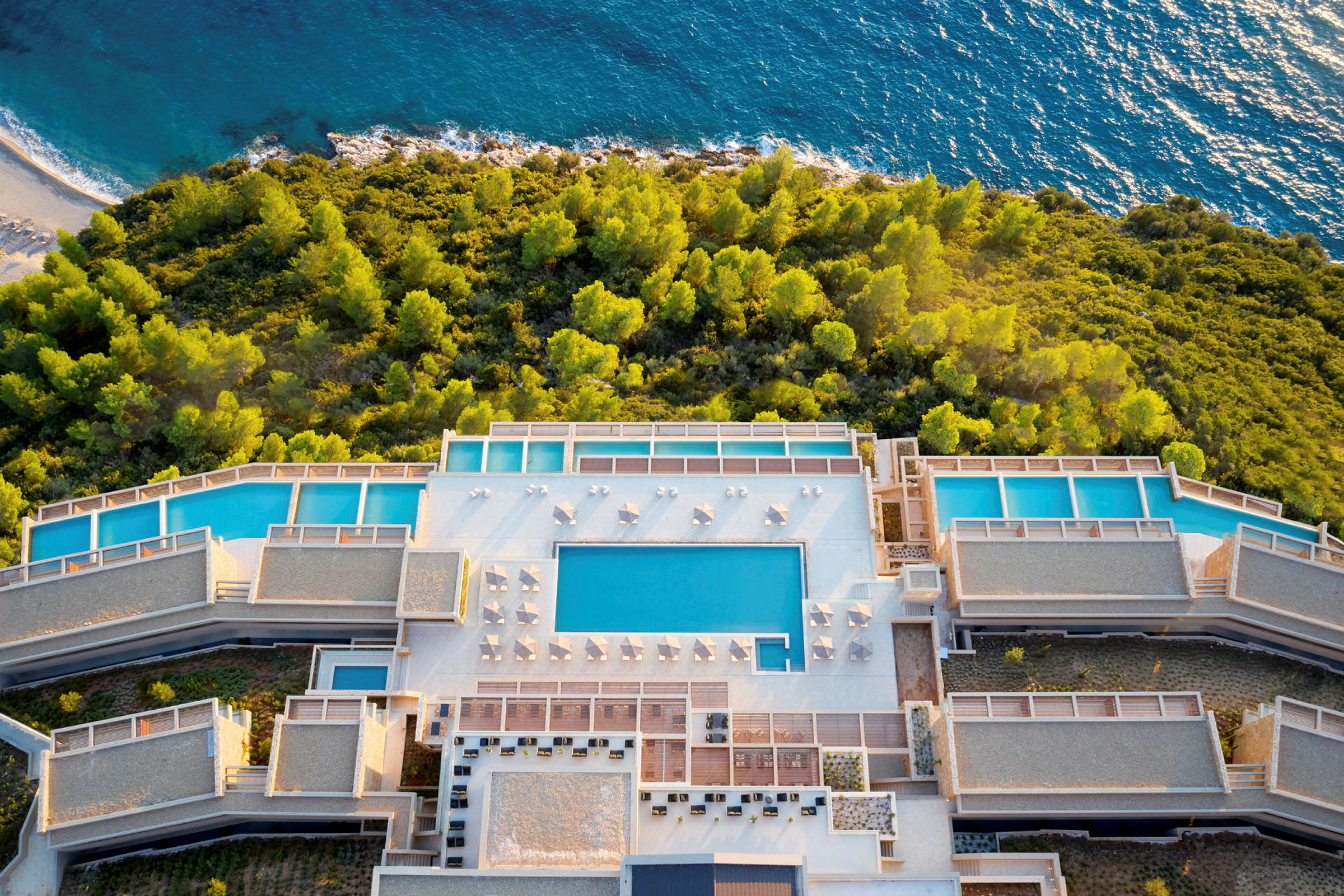 Luftfoto af hotelområde med swimmingpools omgivet af grønne områder tæt på havet. (AI-genereret tekst)