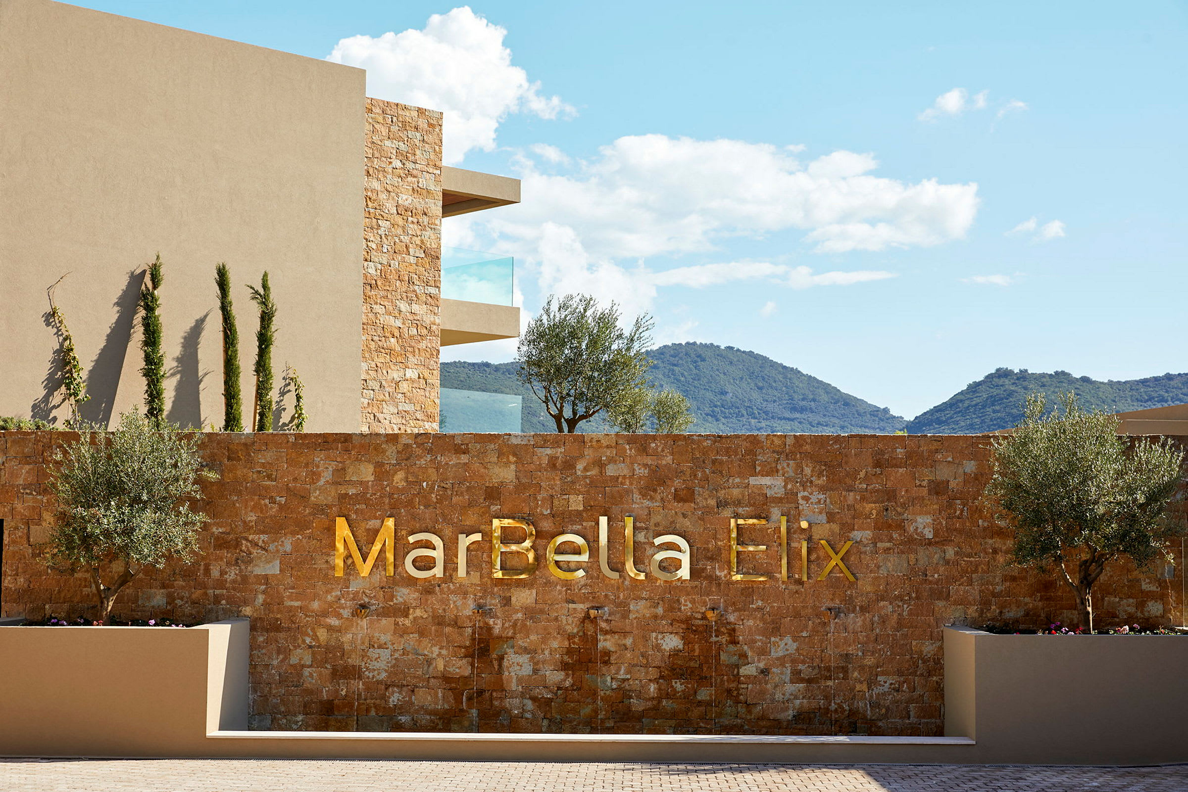 Hotellets facade med teksten 'MarBella Elix' på en stenvæg, omgivet af træer og bjerge i baggrunden. (AI-genereret tekst)