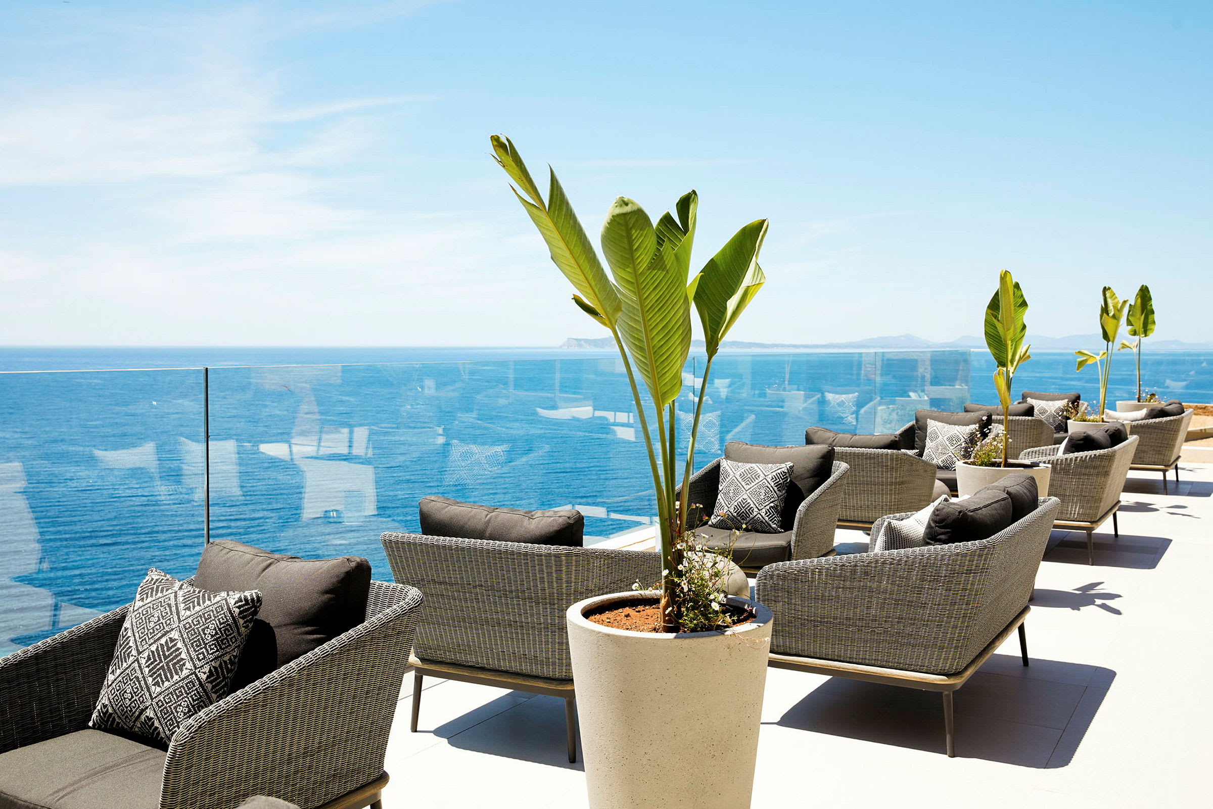 Loungeområde med rattanstole, store potteplanter og udsigt over havet. (AI-genereret tekst)