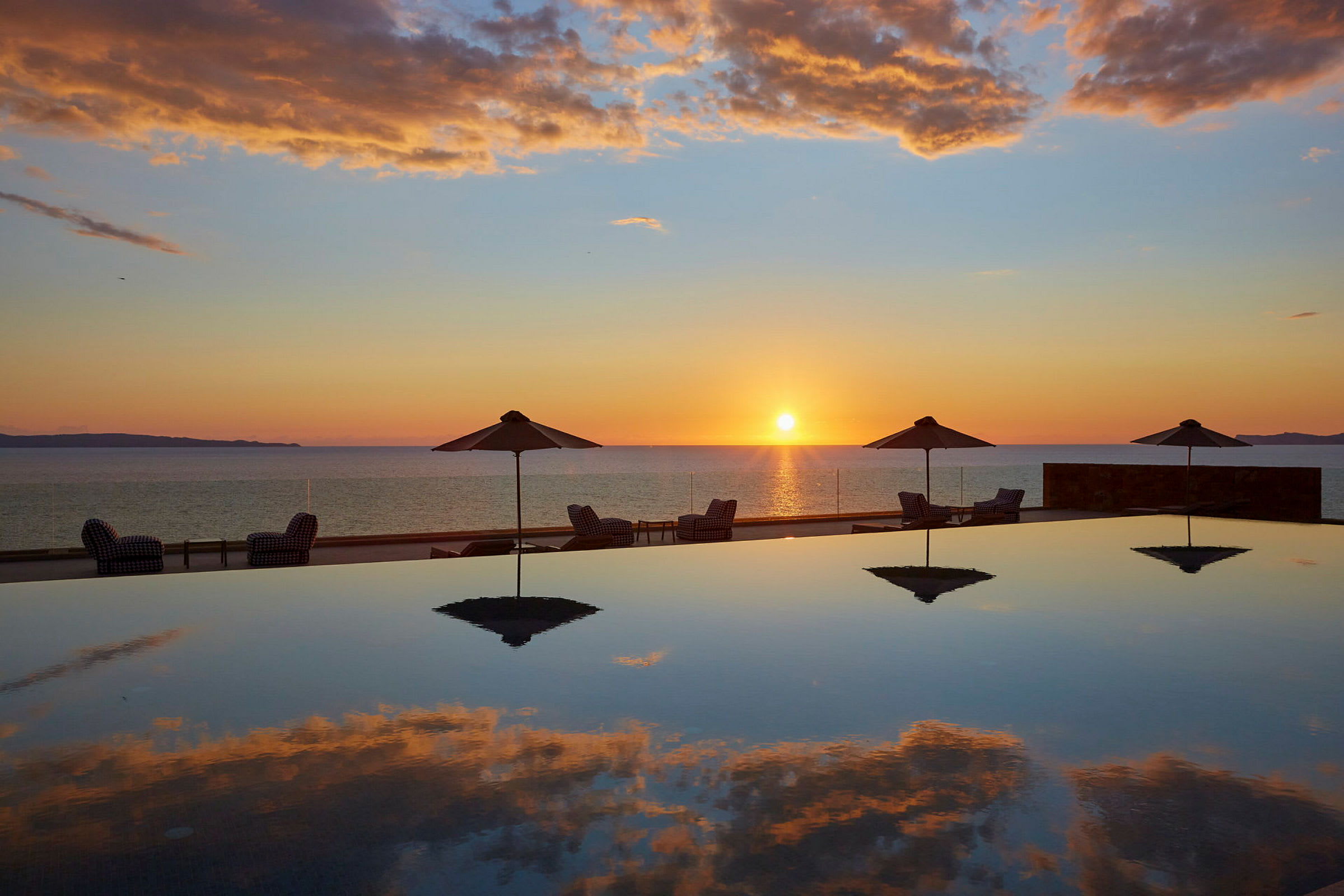 Solnedgang over havet med parasoller og liggestole ved en pool. (AI-genereret tekst)
