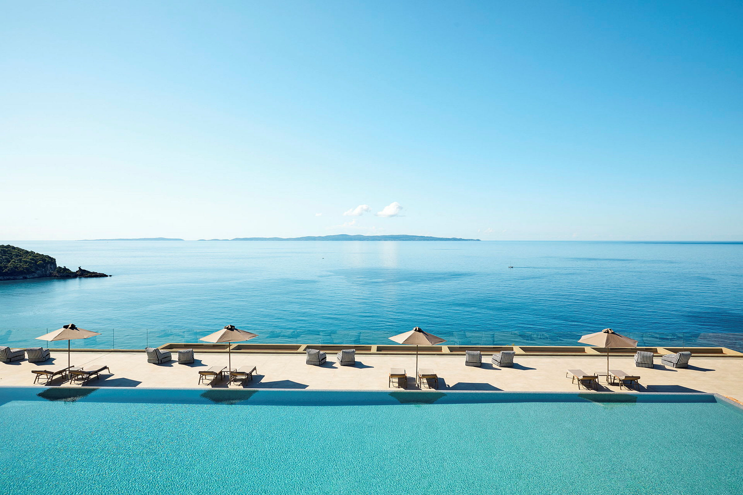 Billede med en infinity-pool med liggestole og parasoller ved udsigt over havet. (AI-genereret tekst)