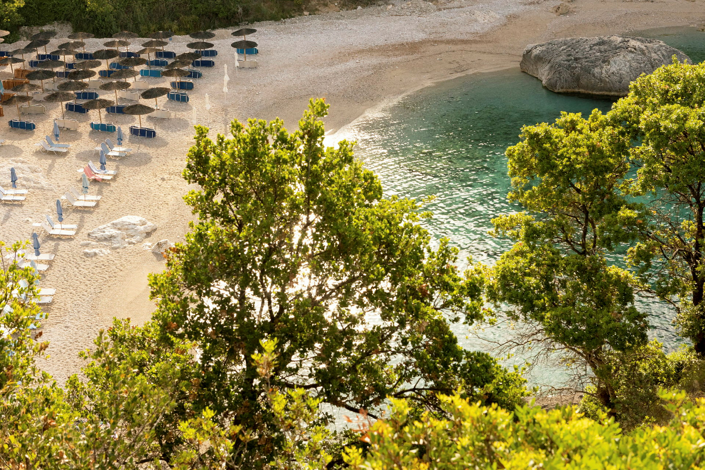 Strand med parasoller, solsenge, vegetation og klart blåt vand. (AI-genereret tekst)