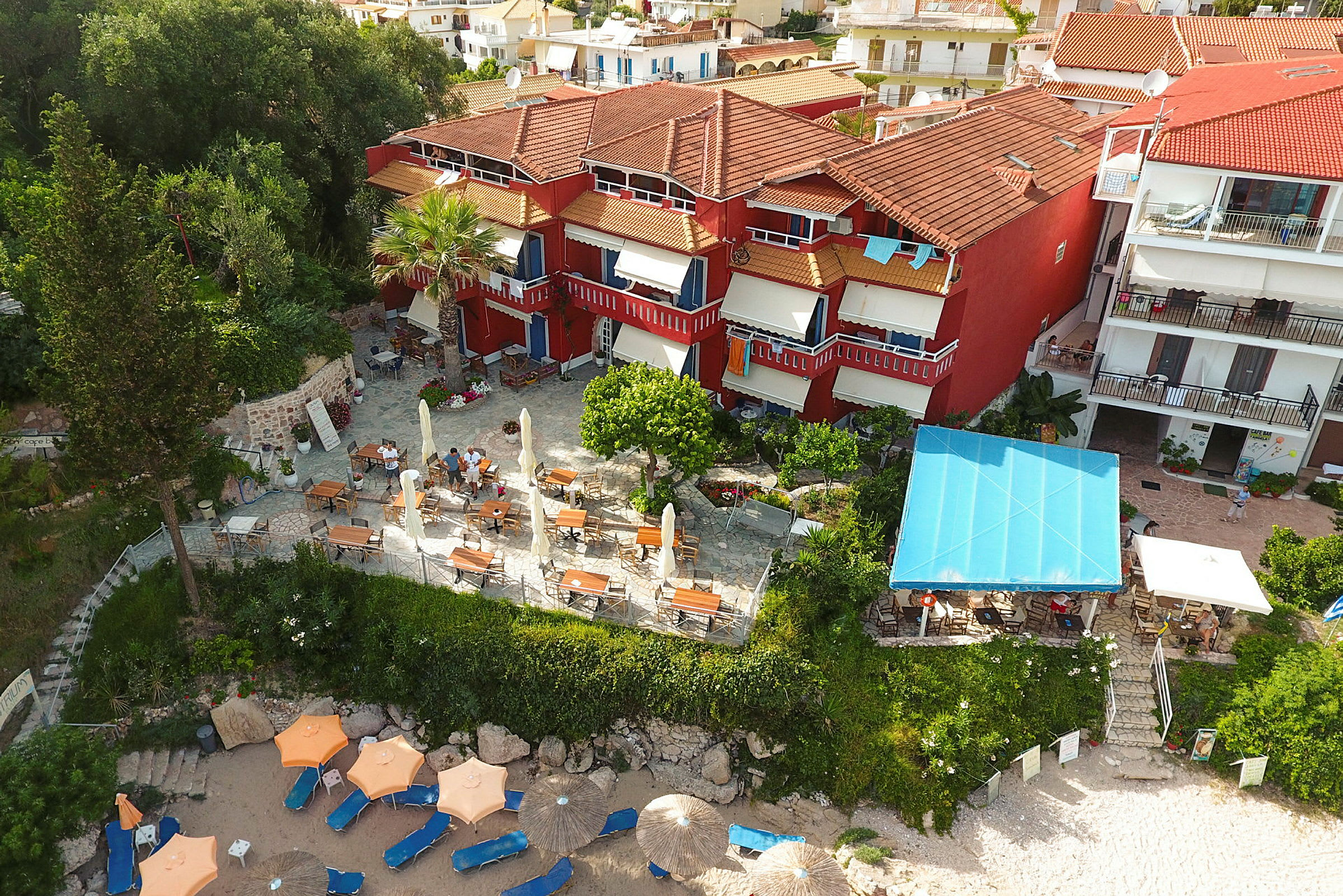 Luftfoto af hotel med røde facader, en terrasse med borde og stole samt parasoller på en sandstrand. (AI-genereret tekst)