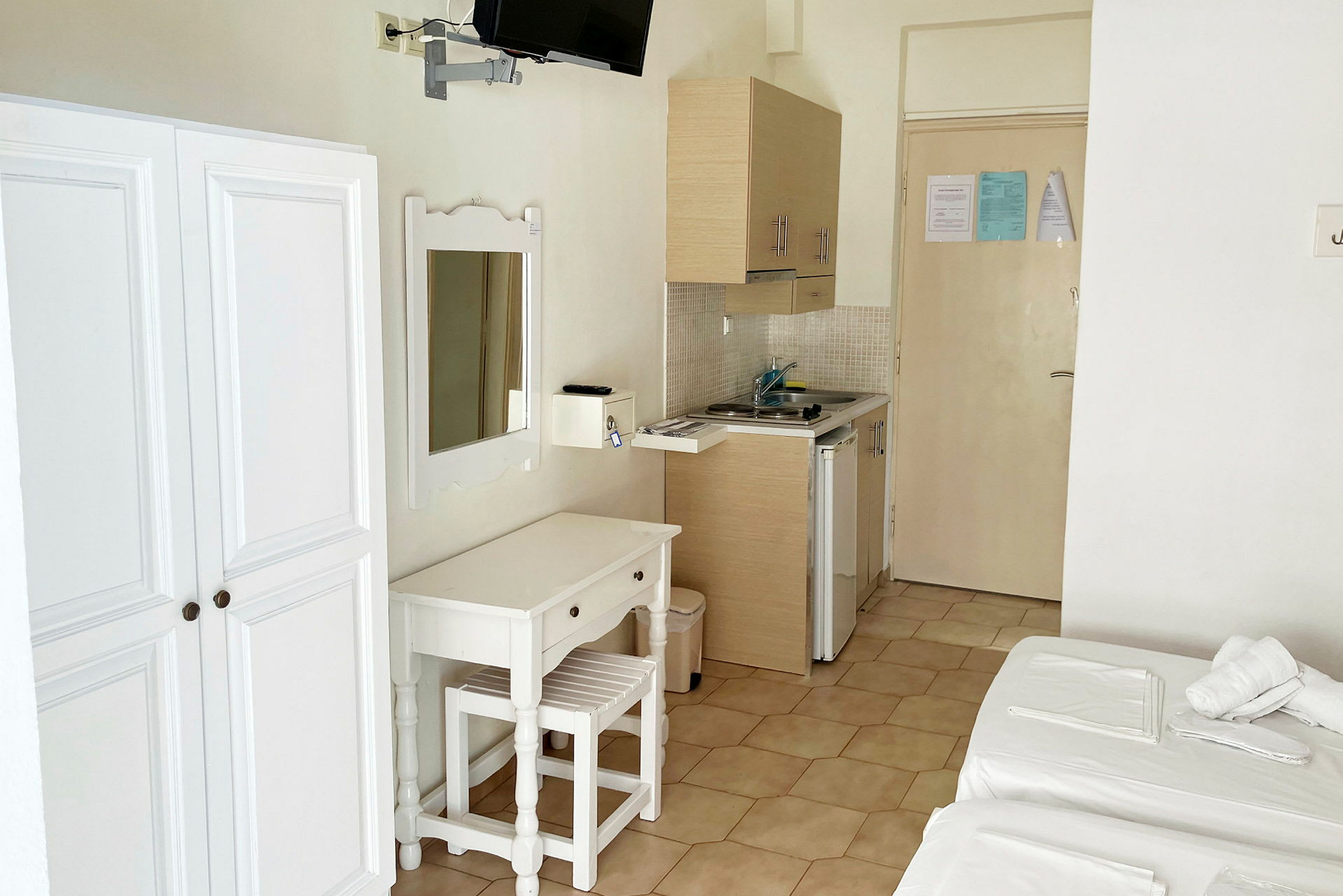 Billede af et hotelværelse med et garderobeskab, et spejl, et skrivebord med stol og en lille køkkensektion med vask. (AI-genereret tekst)