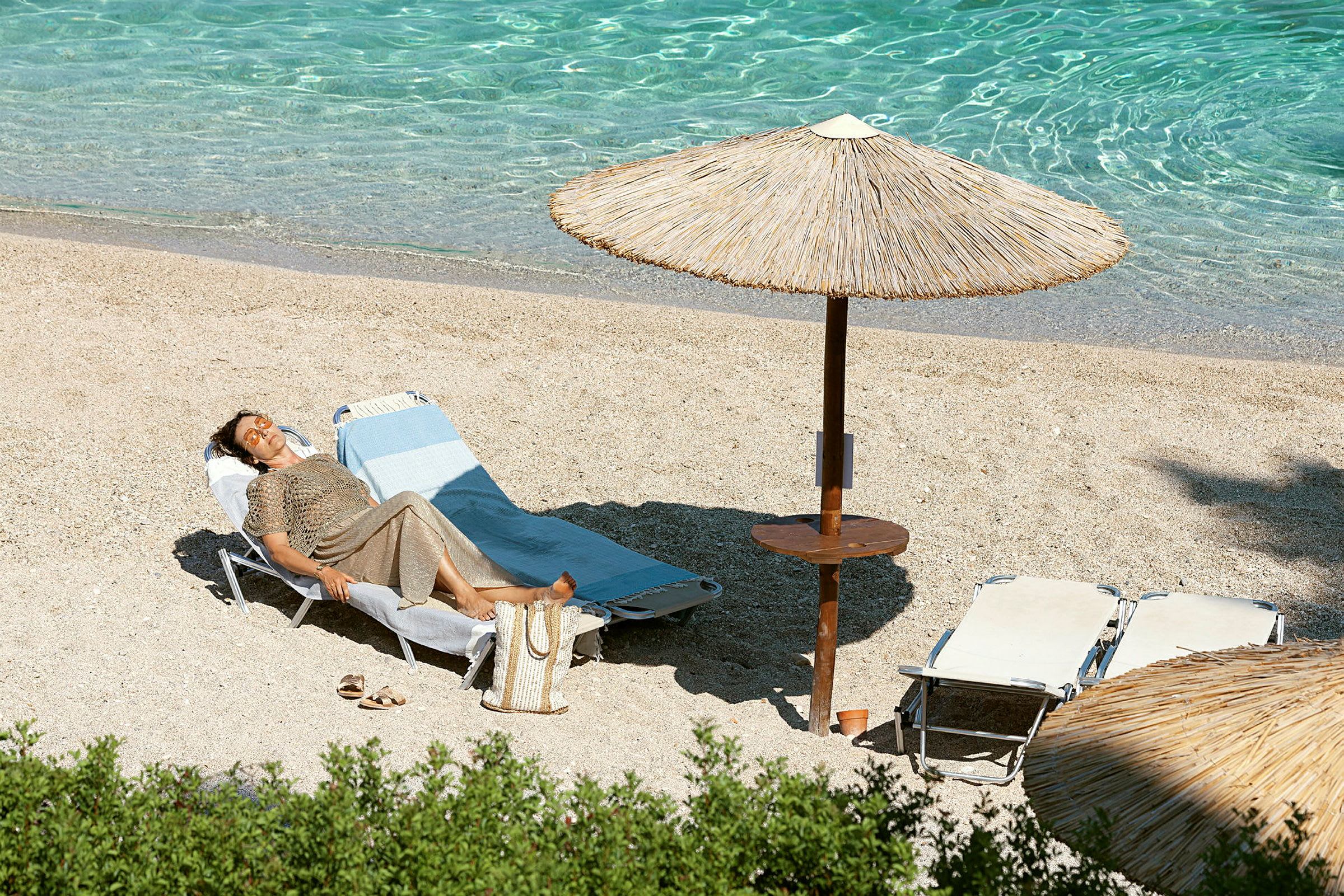 En person ligger på en liggestol på stranden under en parasol ved klart vand. (AI-genereret tekst)