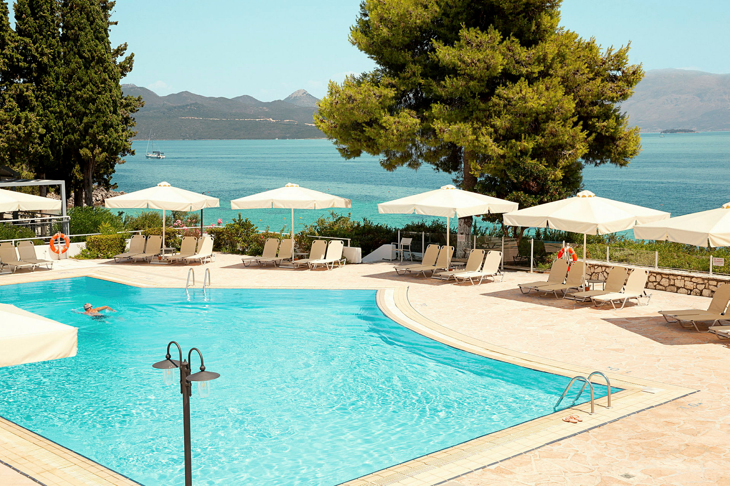 Udendørs swimmingpool med parasoller og liggestole, i baggrunden ses havet og bjergene. (AI-genereret tekst)