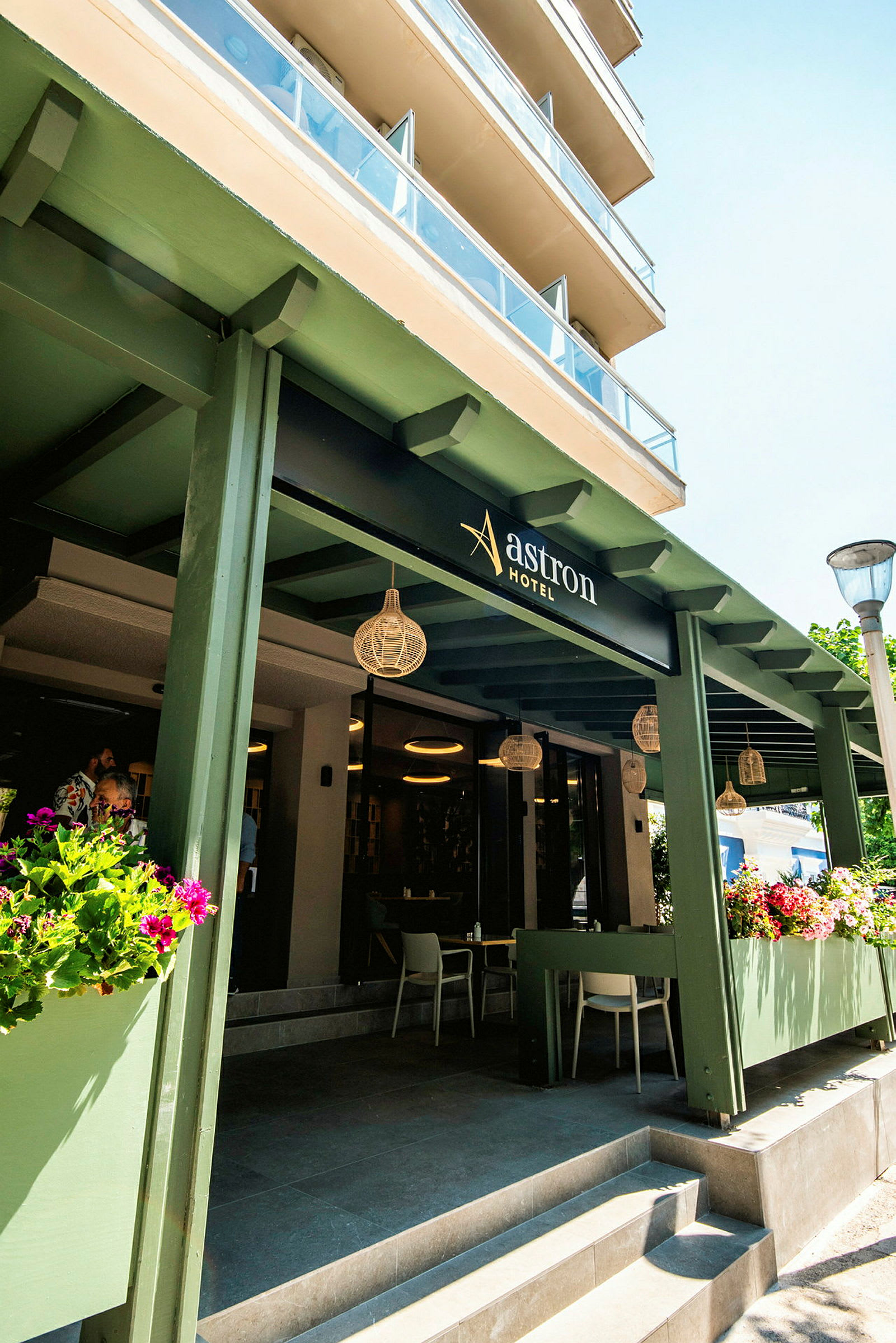 Indgangen til Astron Hotel, med grønne detaljer og blomsterdekorationer. (AI-genereret tekst)