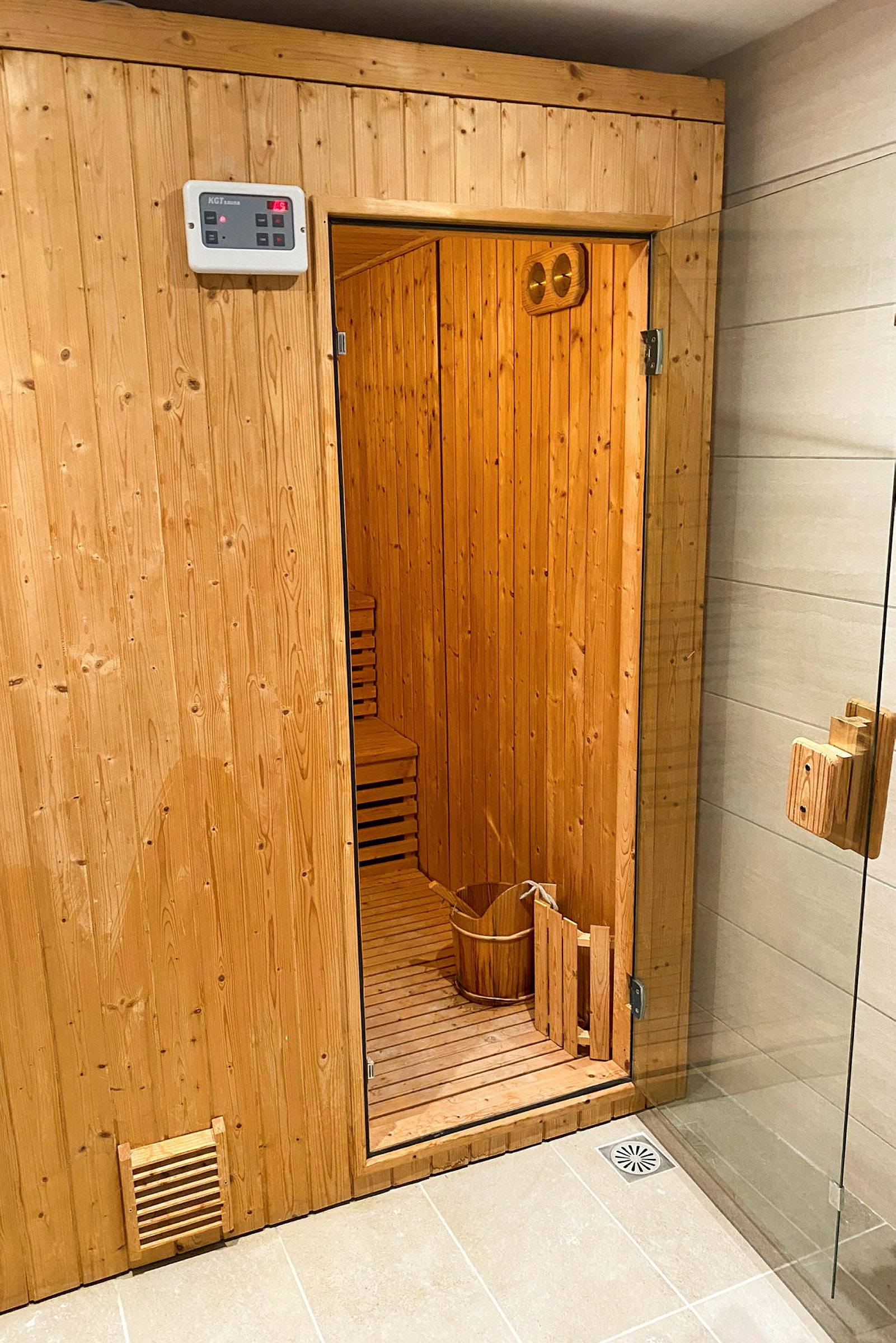 En indendørs sauna i træ med åben dør, en træbalje og gulvafløb synlige. (AI-genereret tekst)