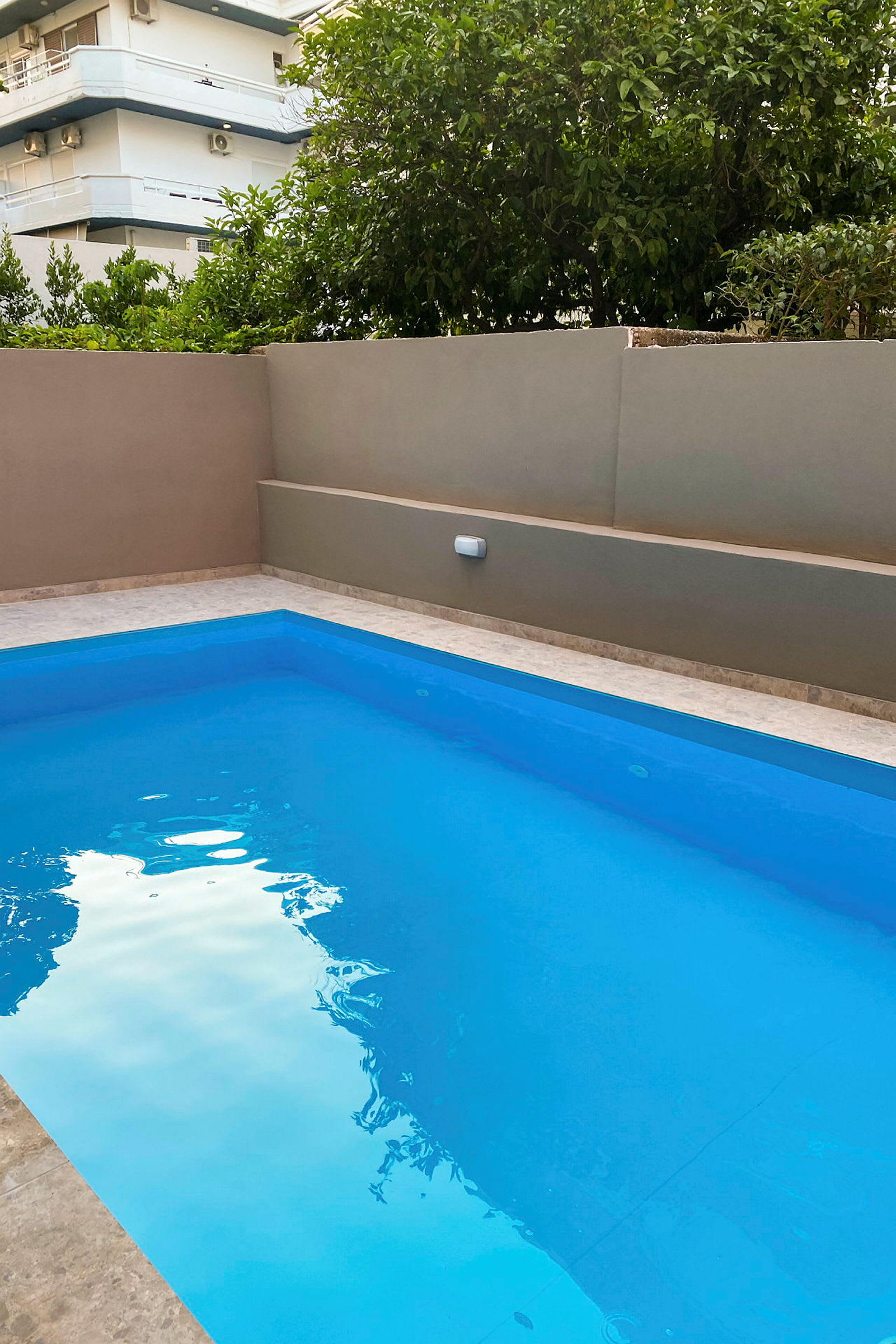 Udendørs pool med klart blåt vand, omgivet af en beige mur og grønt i baggrunden. (AI-genereret tekst)