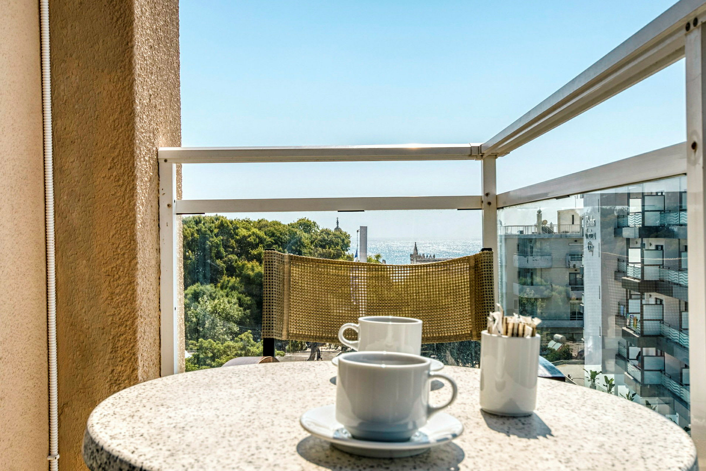 En balkon med udsigt over havet. På bordet er der to kaffekopper og en beholder med sukkerposer. (AI-genereret tekst)