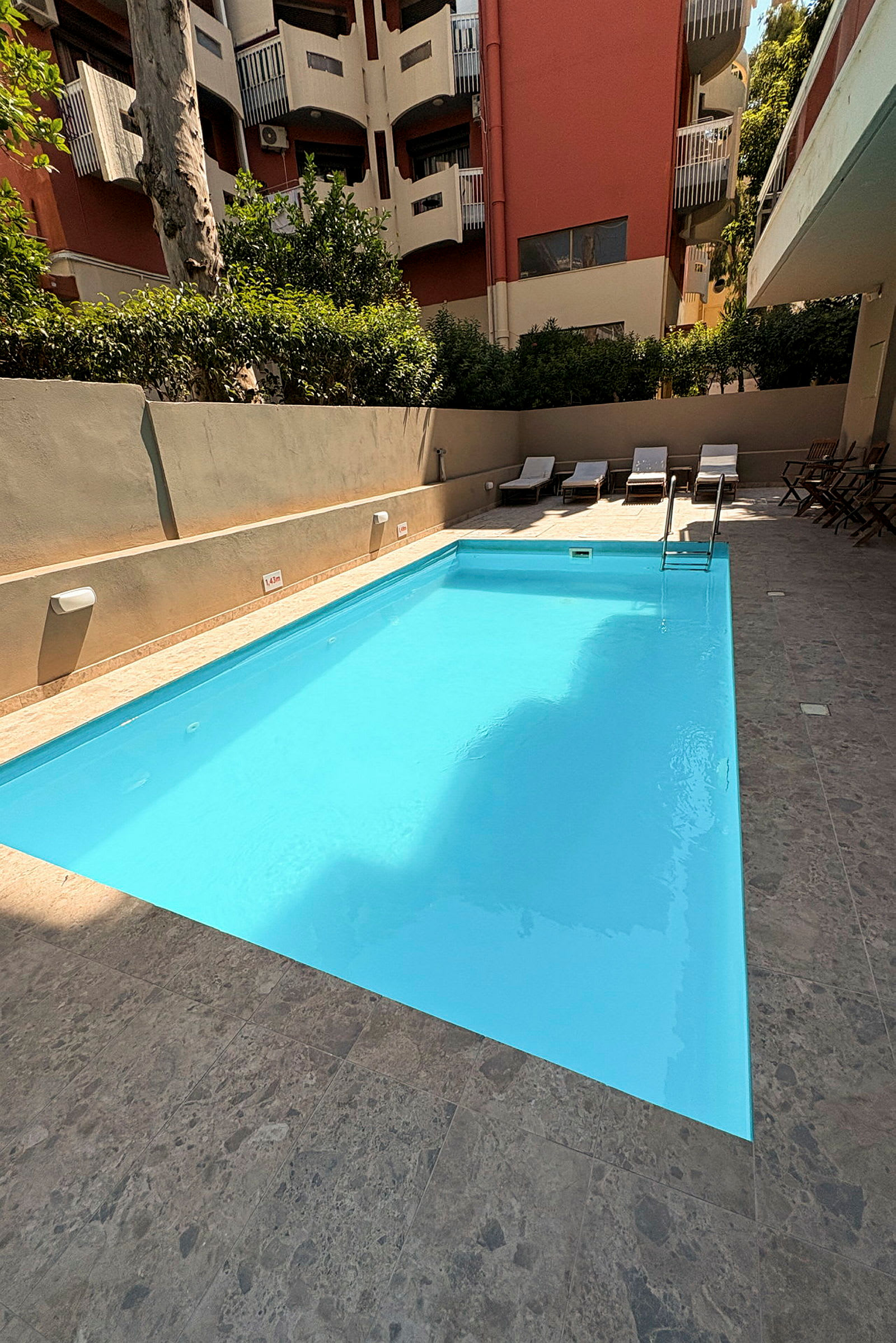 En udendørs swimmingpool omgivet af et stenbelagt område. I baggrunden ses liggestole og grønne planter. (AI-genereret tekst)