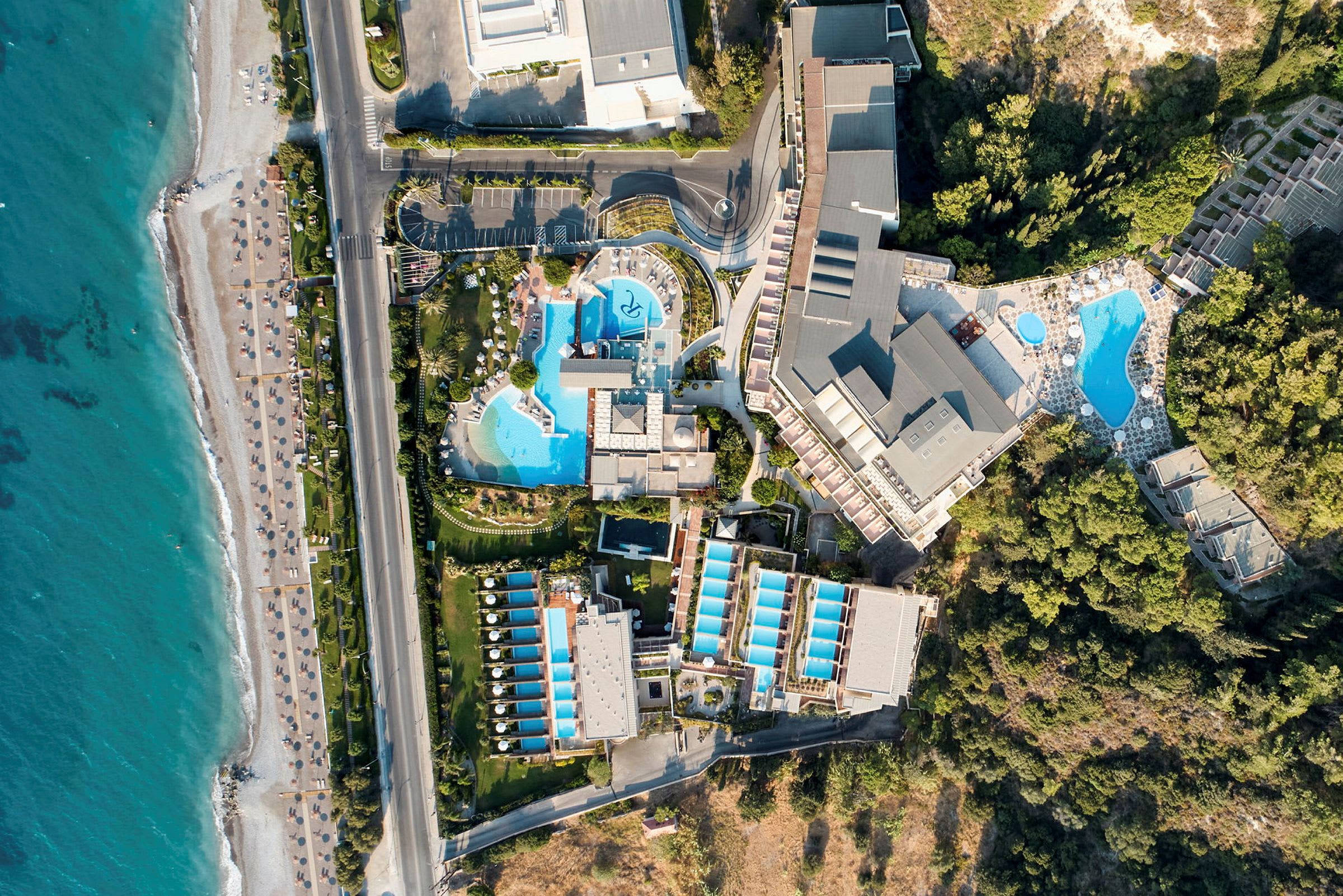 Luftfoto af et hotelområde med pools, bygninger og nærhed til stranden. (AI-genereret tekst)