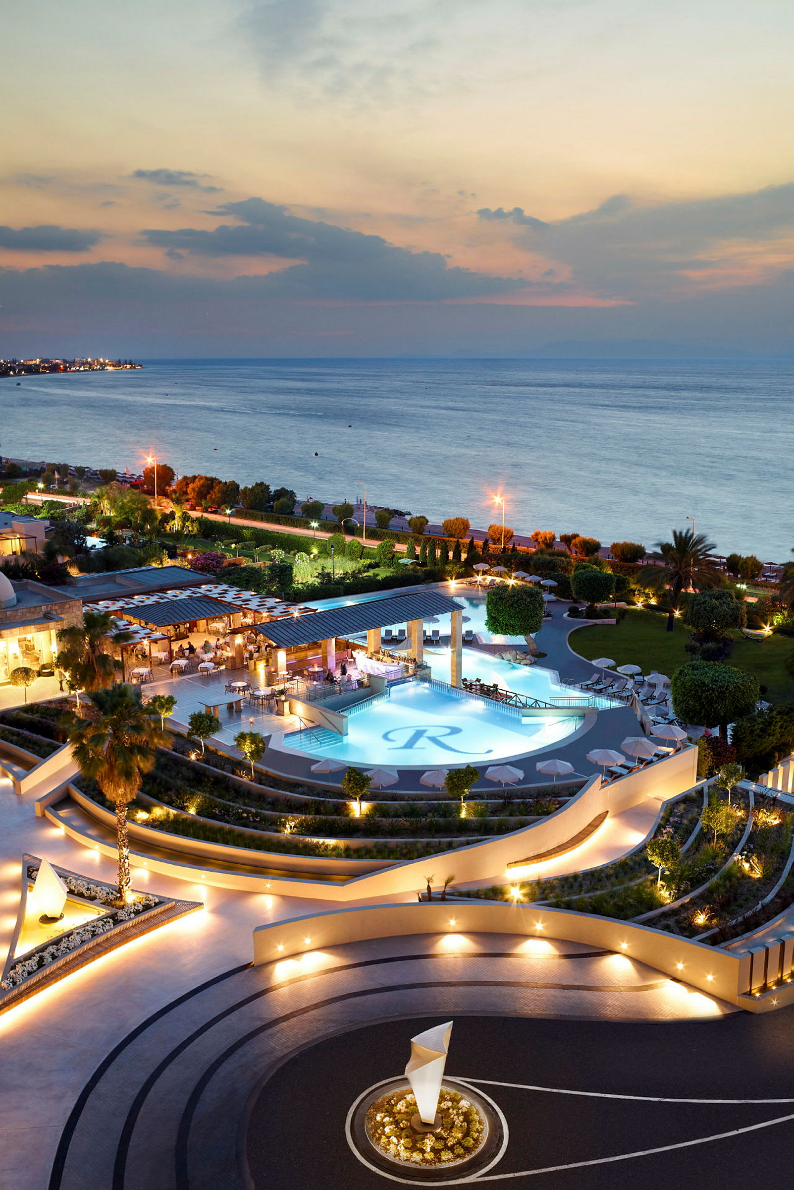 Billede af en elegant resort nær havet med en oplyst pool og omkringliggende haveområder ved solnedgang. (AI-genereret tekst)
