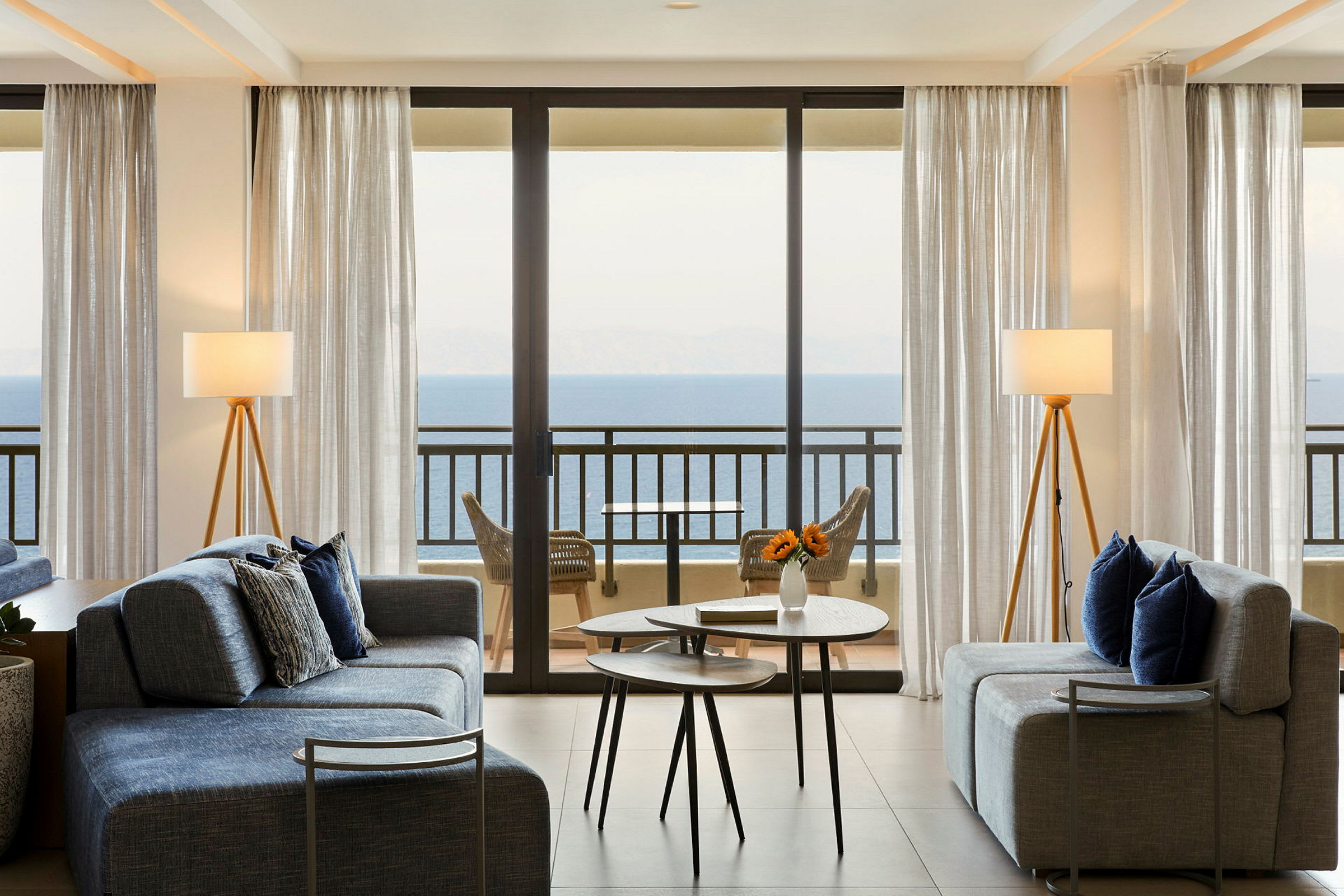 Stue med sofaer og borde, store glasvinduer med udsigt til havet. (AI-genereret tekst)