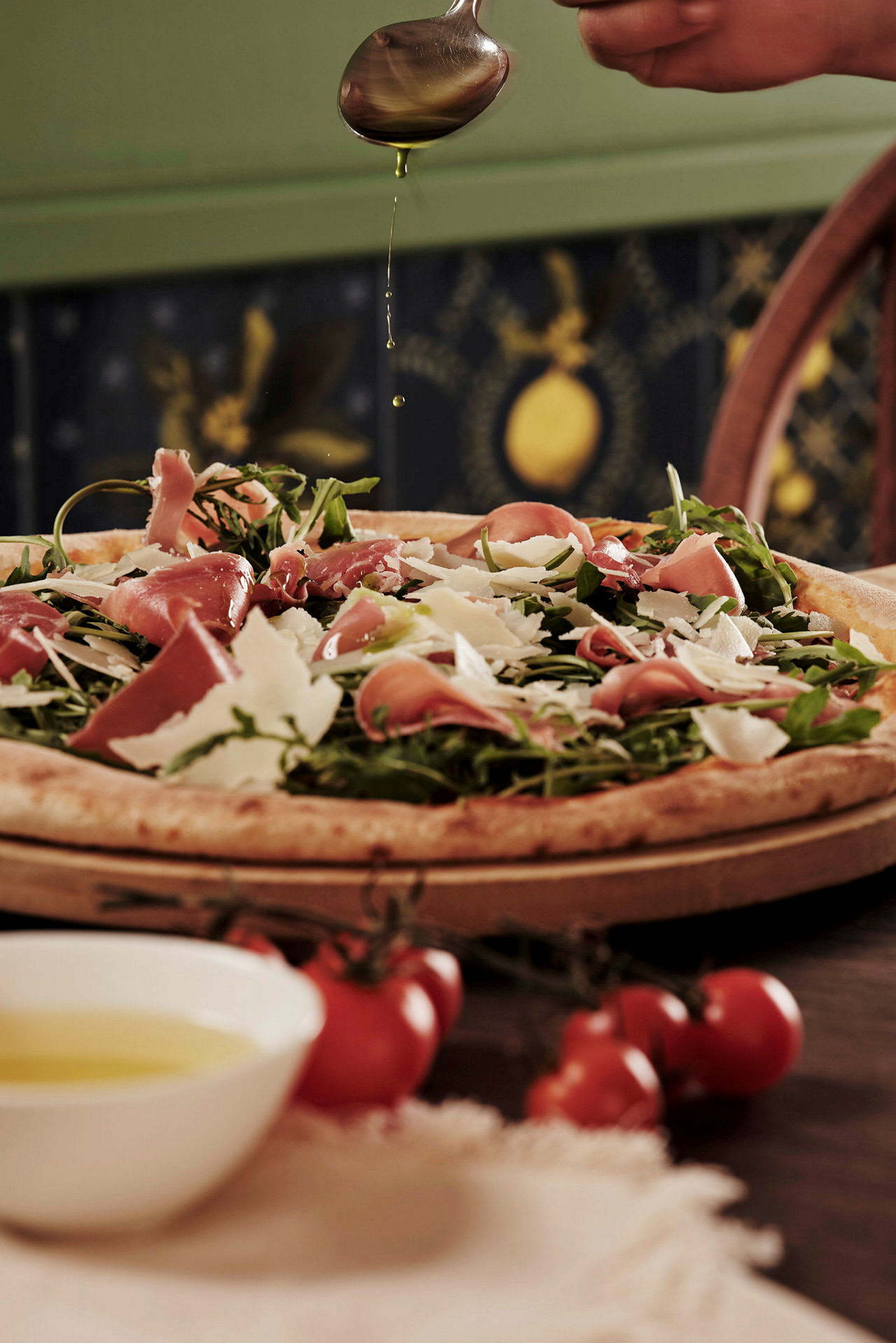 En pizza toppet med skinke, rucola og parmesan, en hånd drypper olivenolie over den. (AI-genereret tekst)