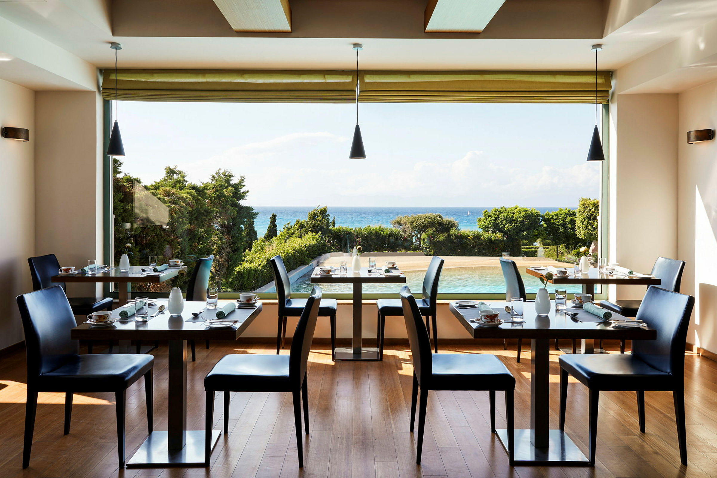 Et lyst restaurantlokale med opdækkede borde og stole, med udsigt over en pool, grønne træer og havet i baggrunden. (AI-genereret tekst)