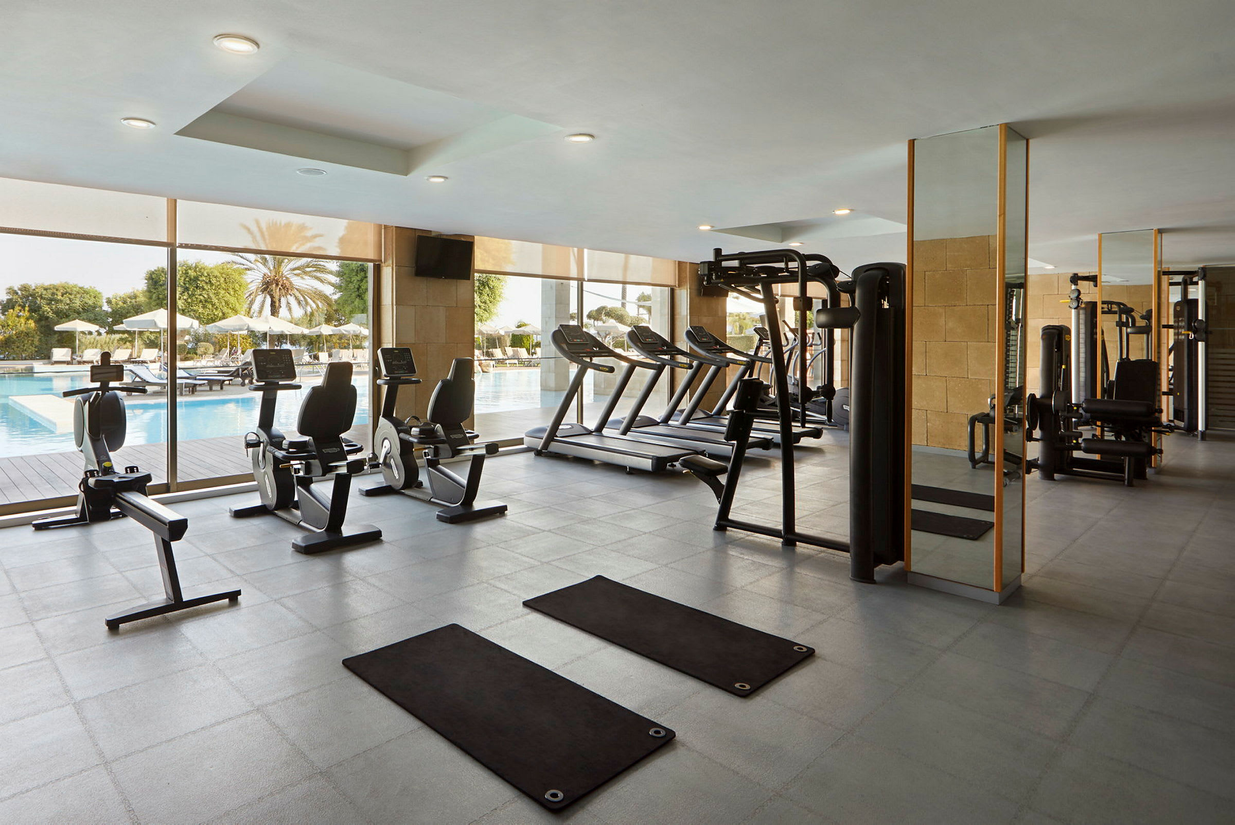 Et fitnesscenter med træningsudstyr og vinduer ud mod et poolområde. (AI-genereret tekst)