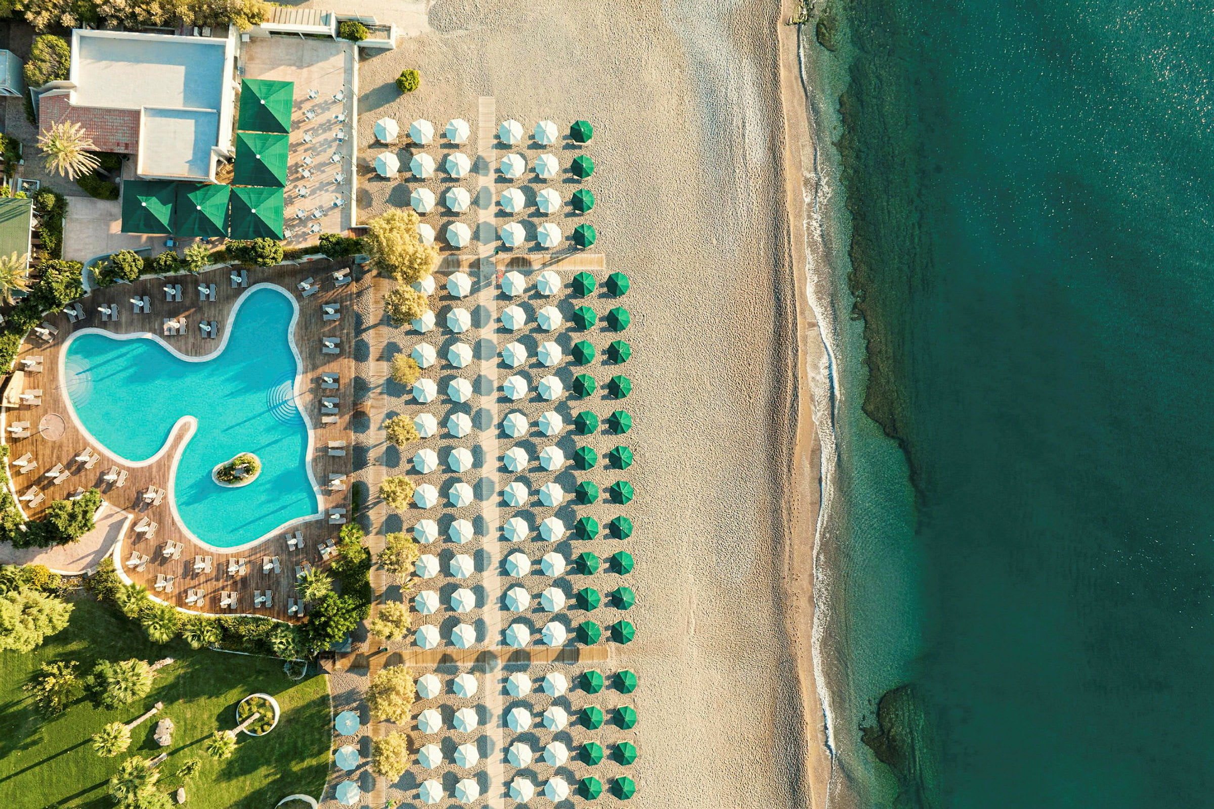 Luftfoto af en hotelpool med omkringliggende liggestole, ved siden af en strand med parasoller og hav. (AI-genereret tekst)