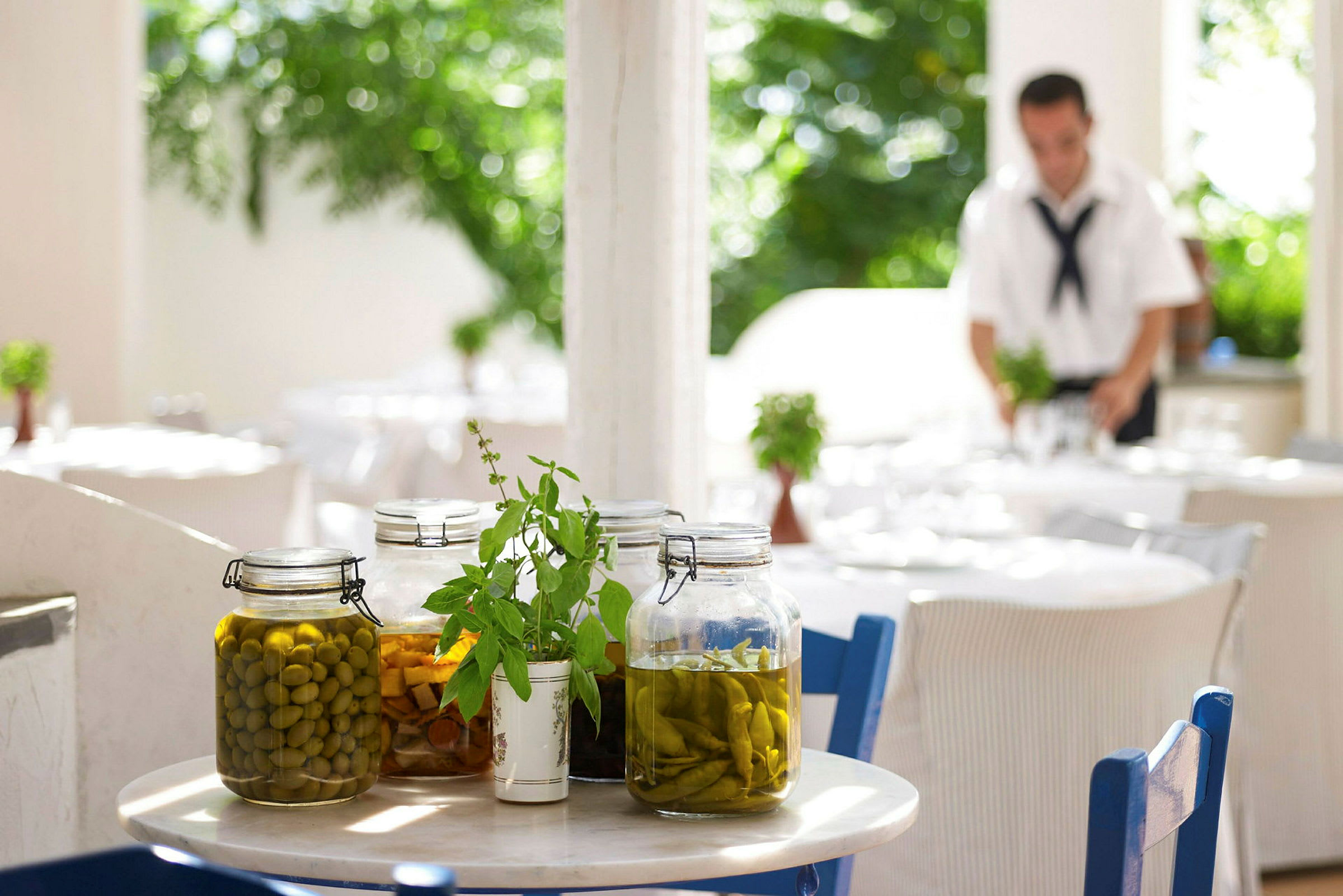 Et bord med glasbeholdere fyldt med syltede grøntsager og dekorationer med urter. Baggrunden viser en udendørsrestaurant. (AI-genereret tekst)