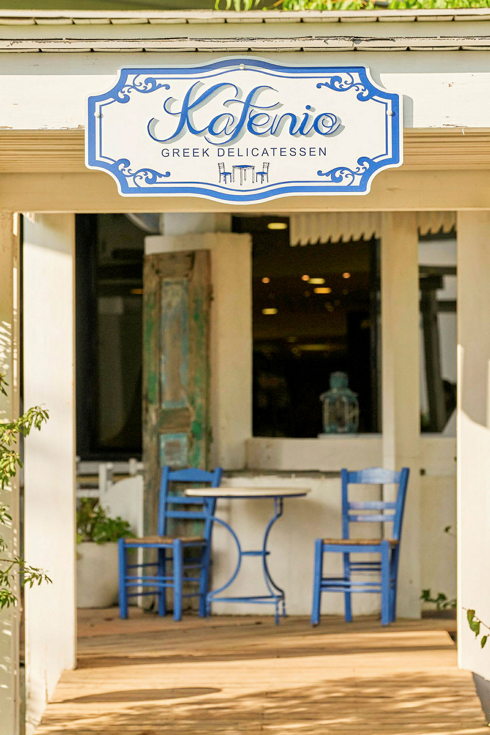 En café med et skilt, der står 'Kafenio', og en gårdhave med blå stole og borde. (AI-genereret tekst)