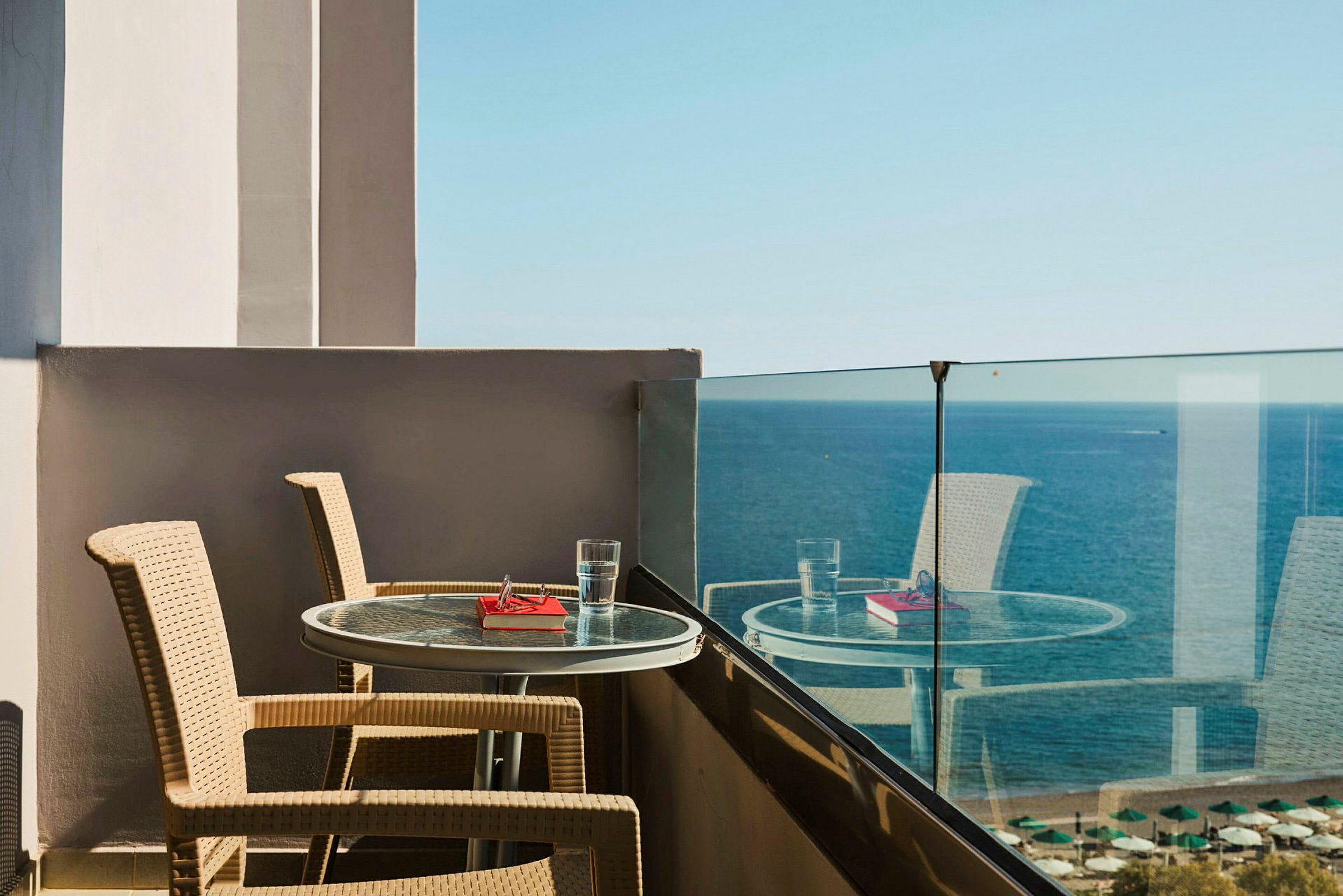 Udsigt fra en balkon med to stole og et bord med et glas vand og en bog foran havet. (AI-genereret tekst)