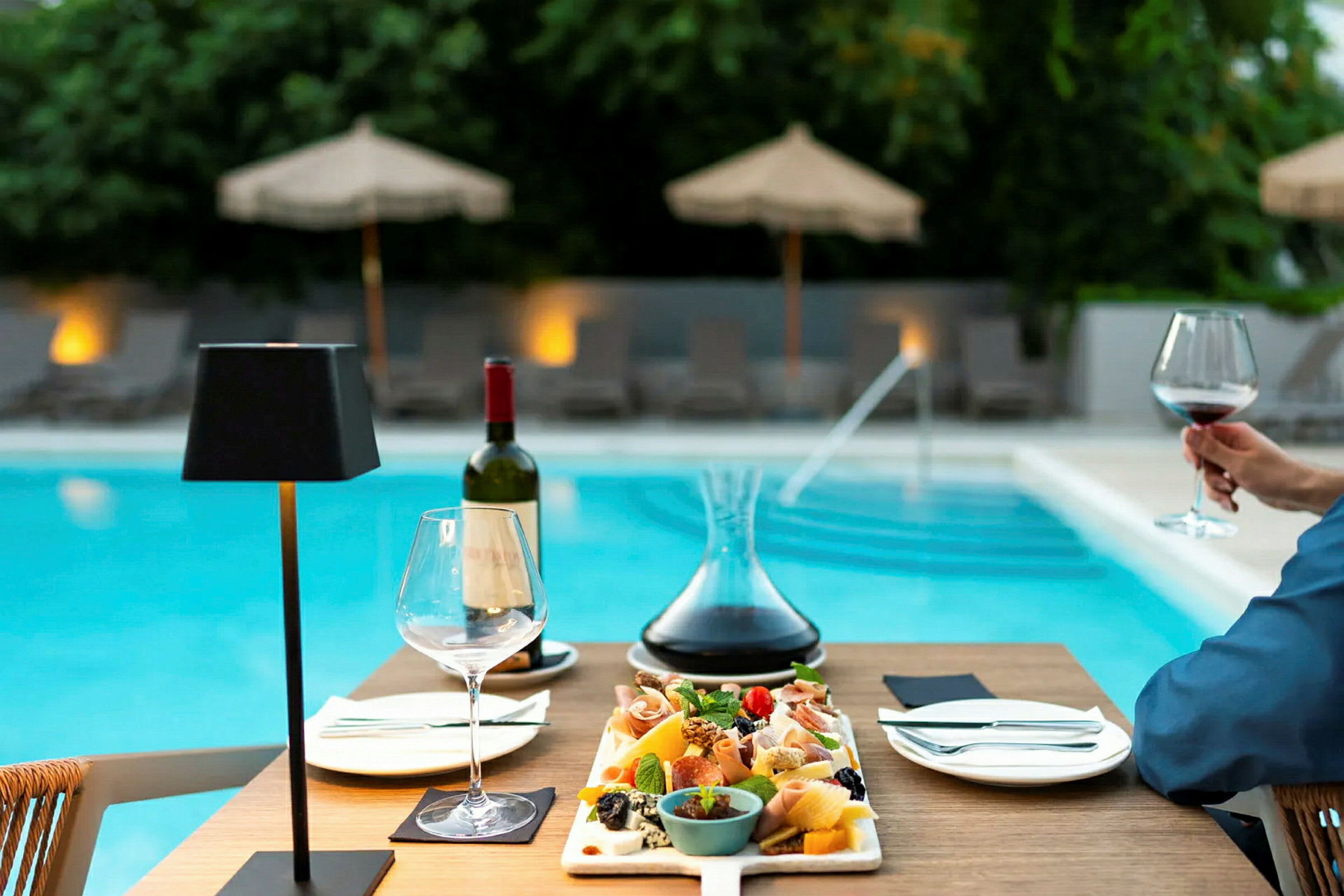 En tallerken med snacks og en karaffel vin på et bord ved en pool. (AI-genereret tekst)
