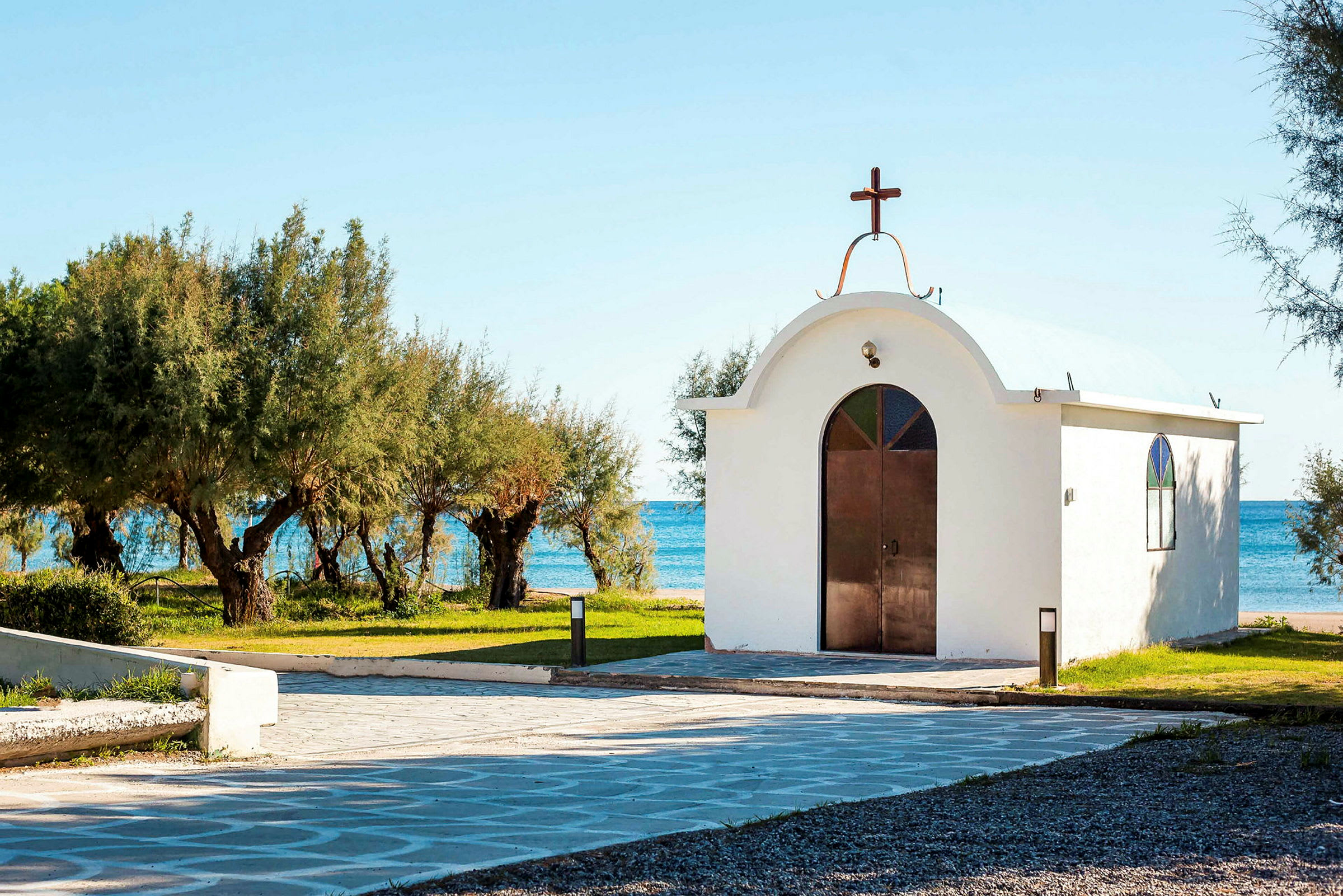 En hvid kirke med kobberkors og glasvinduer, omgivet af grønne træer ved havet. (AI-genereret tekst)