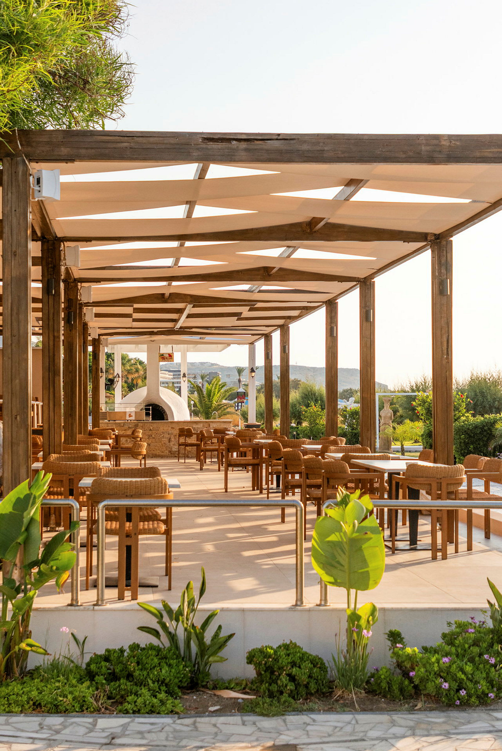 En udendørsrestaurant med træstole og borde under en pergola, omgivet af grønne planter. (AI-genereret tekst)