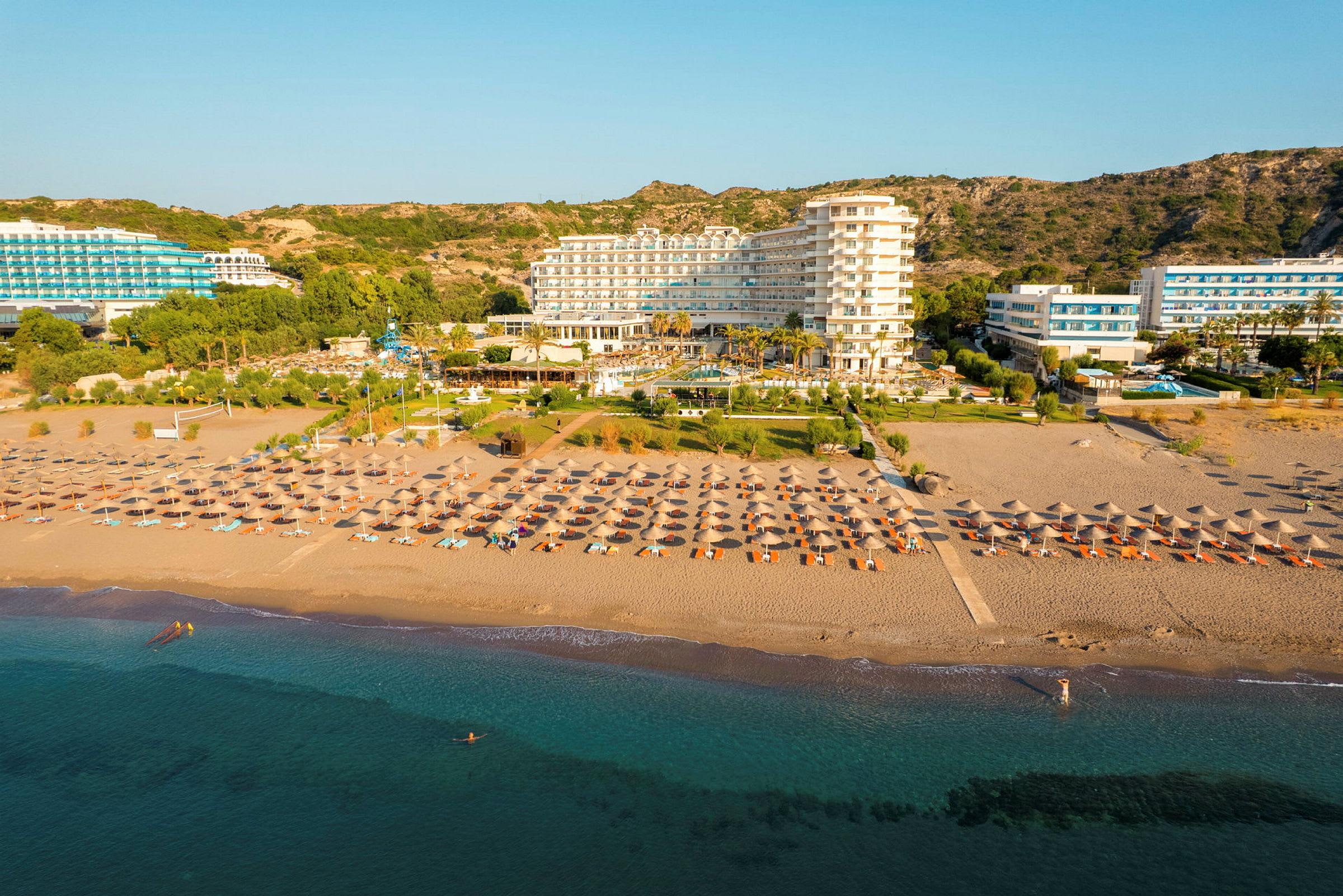 Et hotel ved stranden med liggestole og parasoller foran samt en klar blå himmel i baggrunden. (AI-genereret tekst)