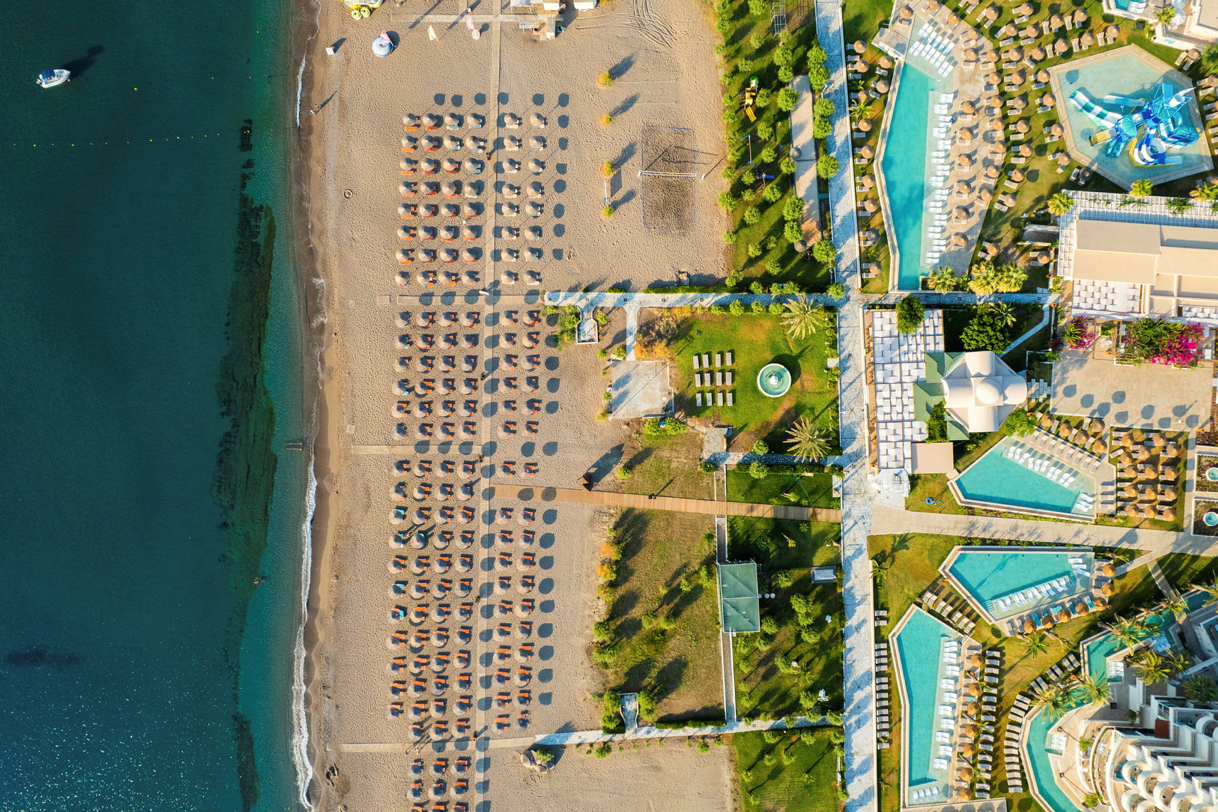Luftfoto af en strand med liggestole og parasoller ved siden af et hotelområde med pools og grønne områder. (AI-genereret tekst)