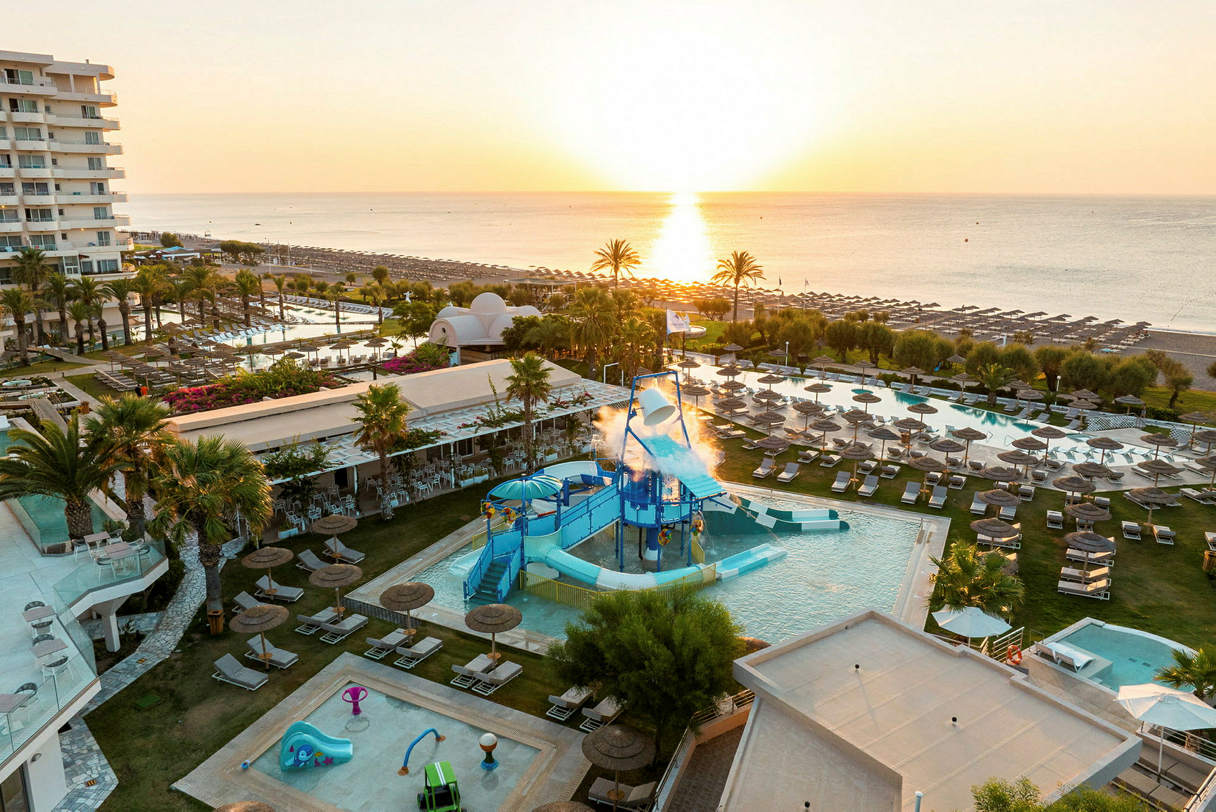 En udsigt over et ferieresort ved stranden med pools og vandlegeområde, omgivet af liggestole og parasoller under en solnedgang. (AI-genereret tekst)