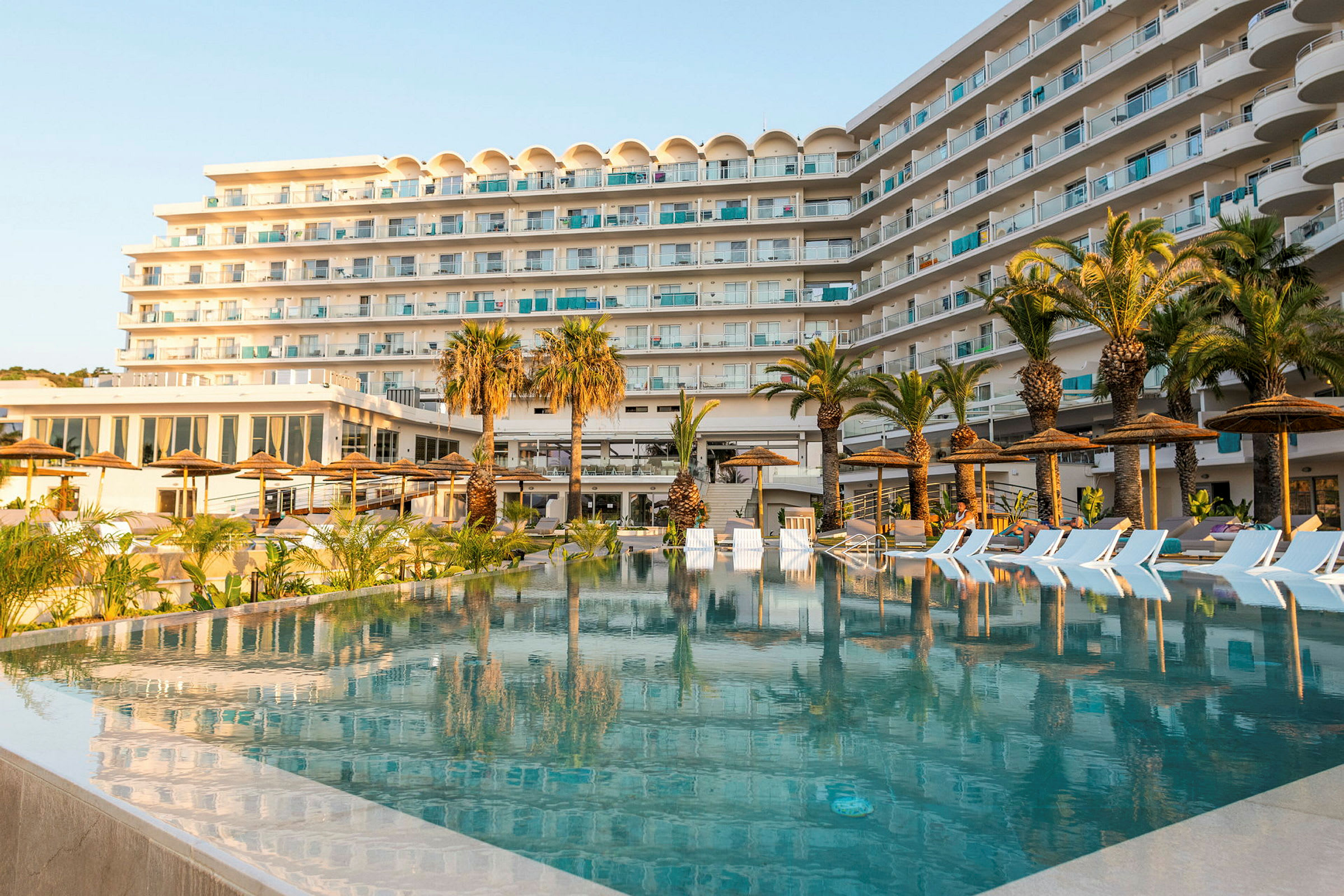 Et hotel med flere etager foran en pool omgivet af palmer og liggestole. (AI-genereret tekst)