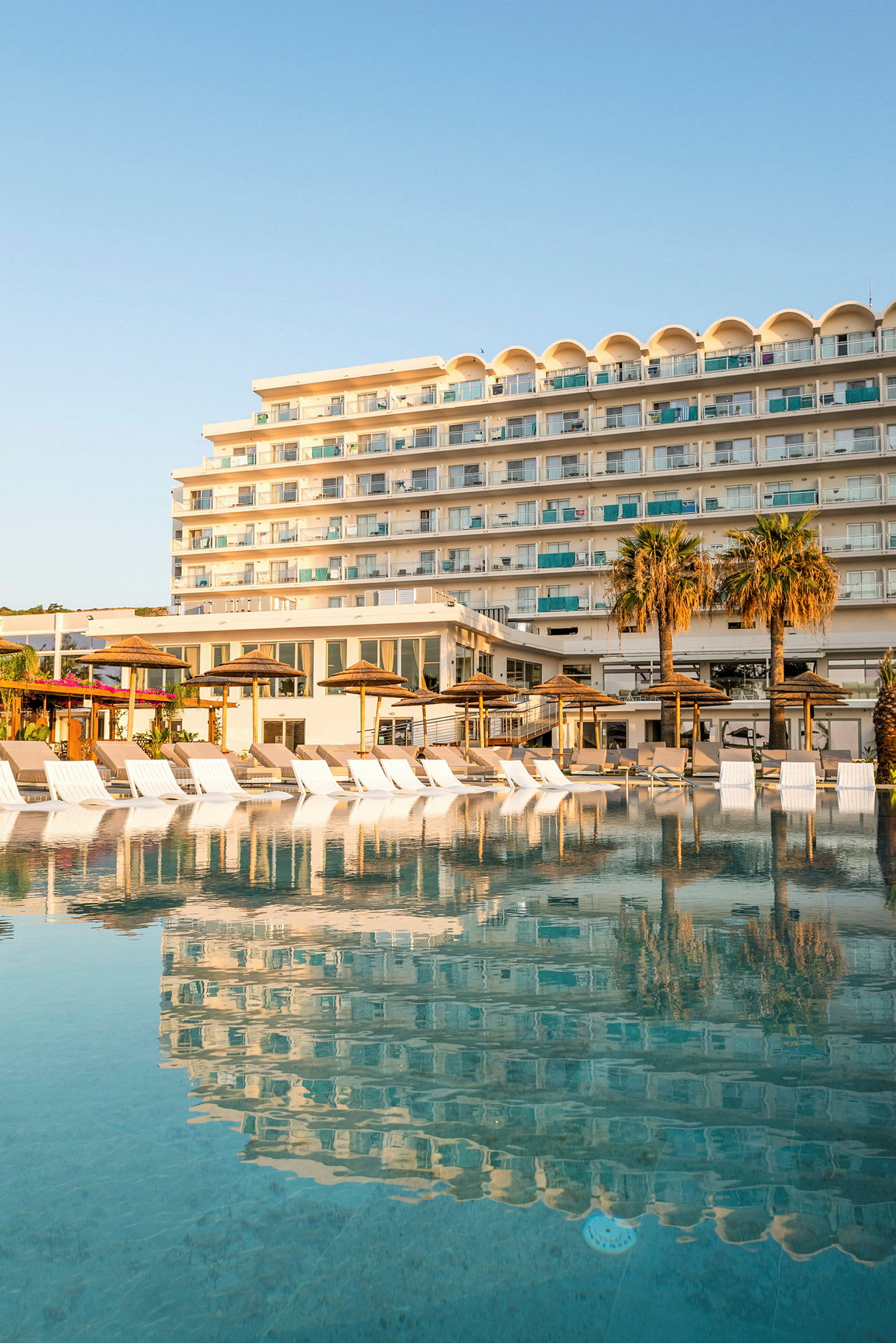 Hotelbygning ved en stor pool med liggestole og parasoller i forgrunden. (AI-genereret tekst)