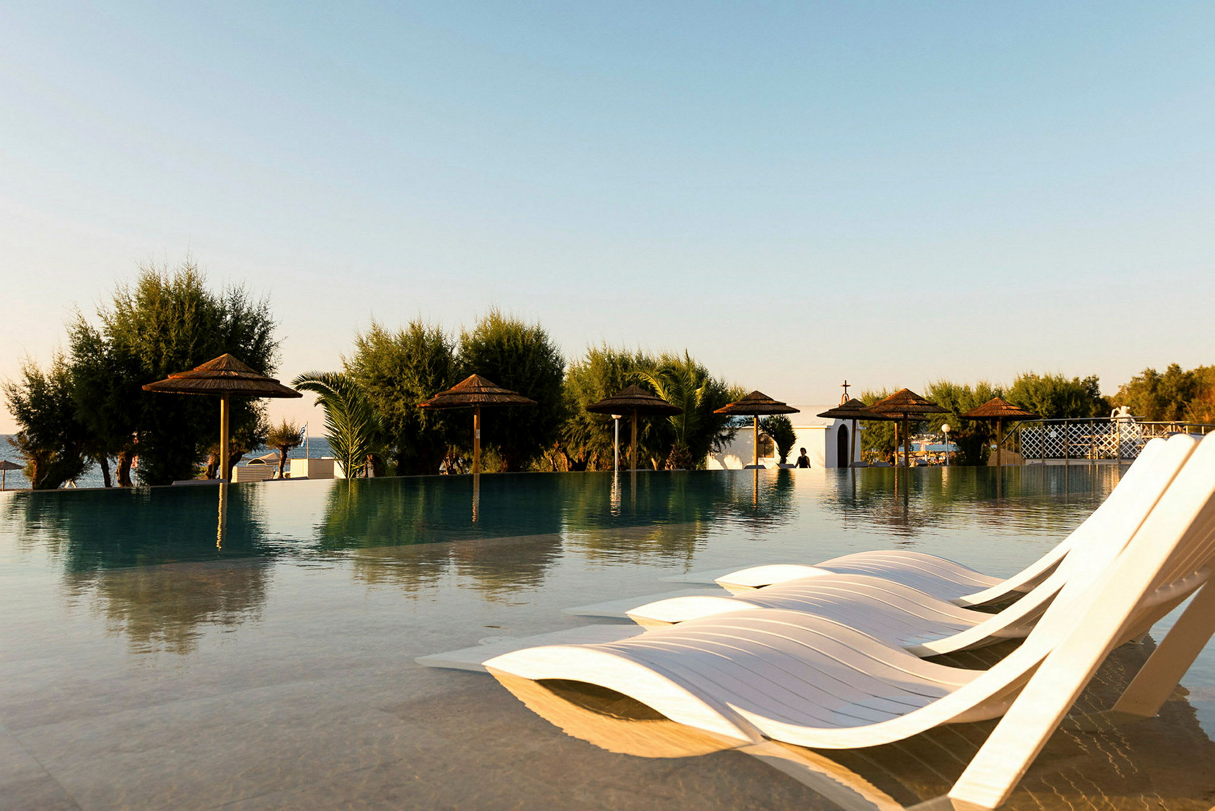 En infinity-pool med liggestole og halmtagdede parasoller omgivet af grønne træer, med et åbent landskab i baggrunden. (AI-genereret tekst)