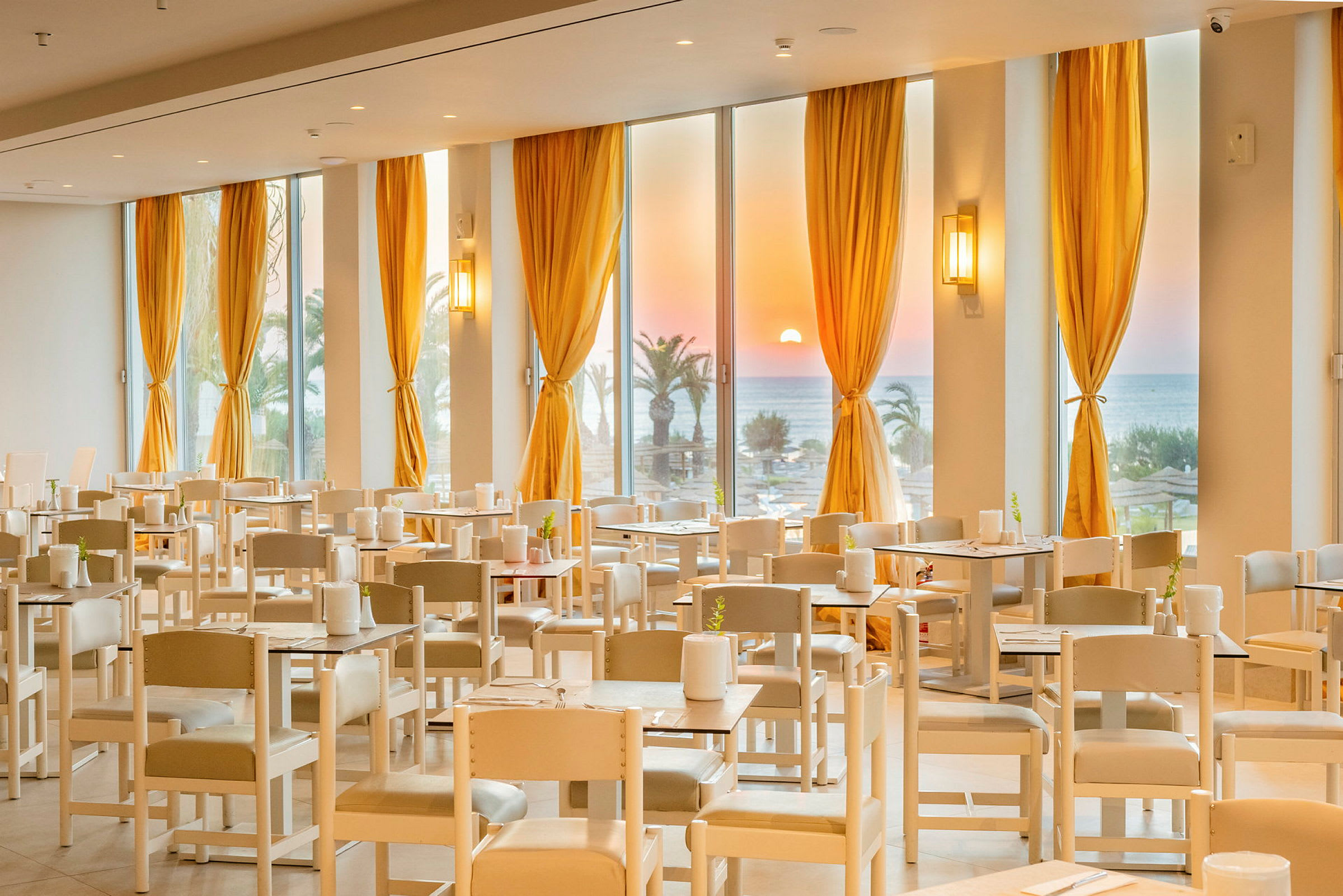En restaurant med bordopstillinger og hvide stole. Store vinduer med gule gardiner viser en solnedgang over havet og palmer. (AI-genereret tekst)