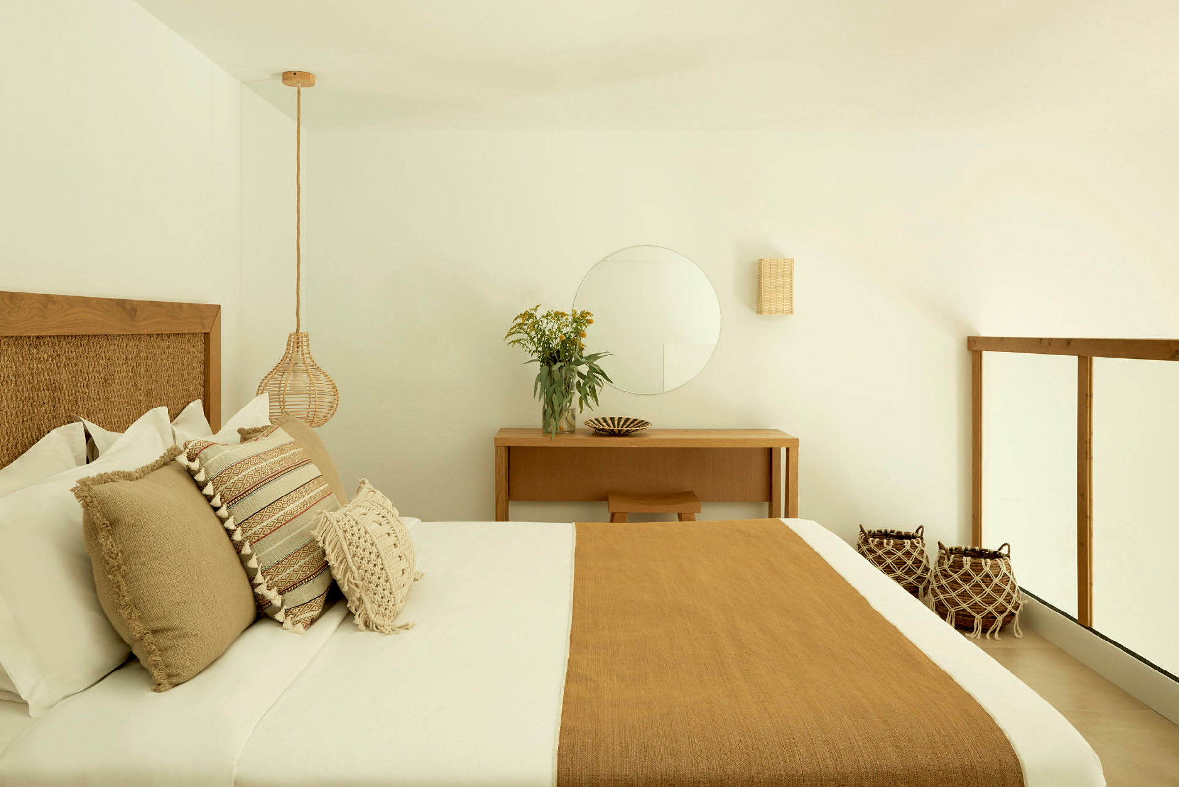 Et elegant soveværelse med en dobbeltseng, dekoreret med brune, beige og hvide puder samt et matchende sengetæppe. Væggene er hvide med minimalistisk indretning og et spejl over et skrivebord. (AI-genereret tekst)