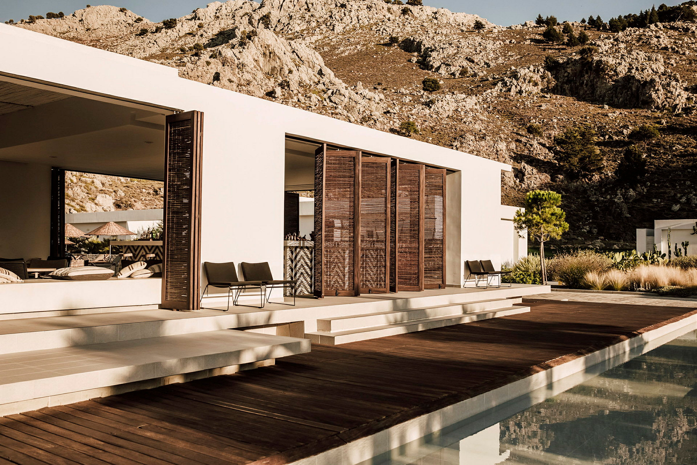 Moderne villa med træterrasse og pool foran bjerglandskab. (AI-genereret tekst)