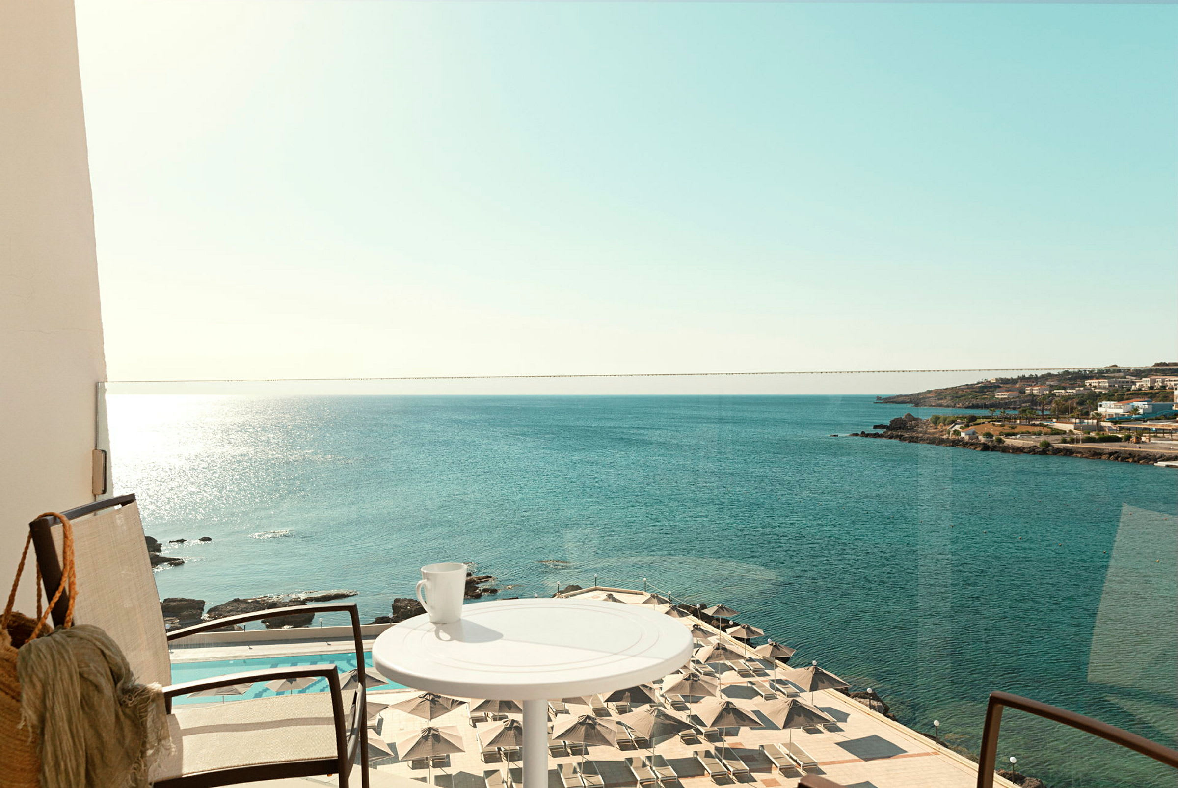 En balkon med udsigt over havet, parasoller ved en pool og en kop på et bord. (AI-genereret tekst)