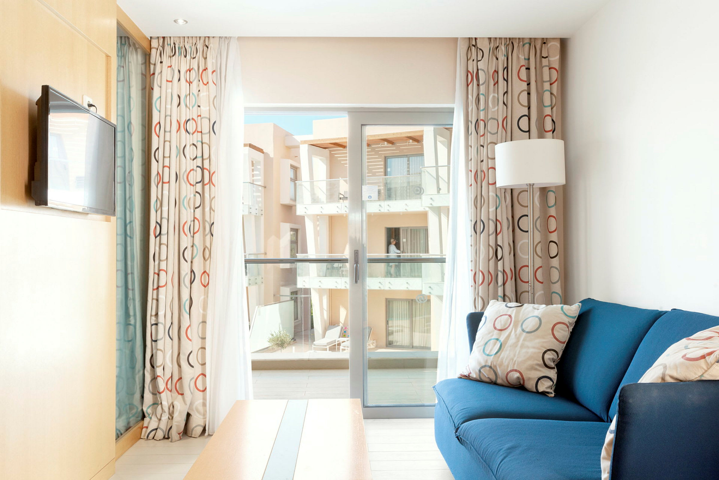 Et lyst hotelværelse med blå sofa, gardiner med cirkelmønster og adgang til en altan. (AI-genereret tekst)
