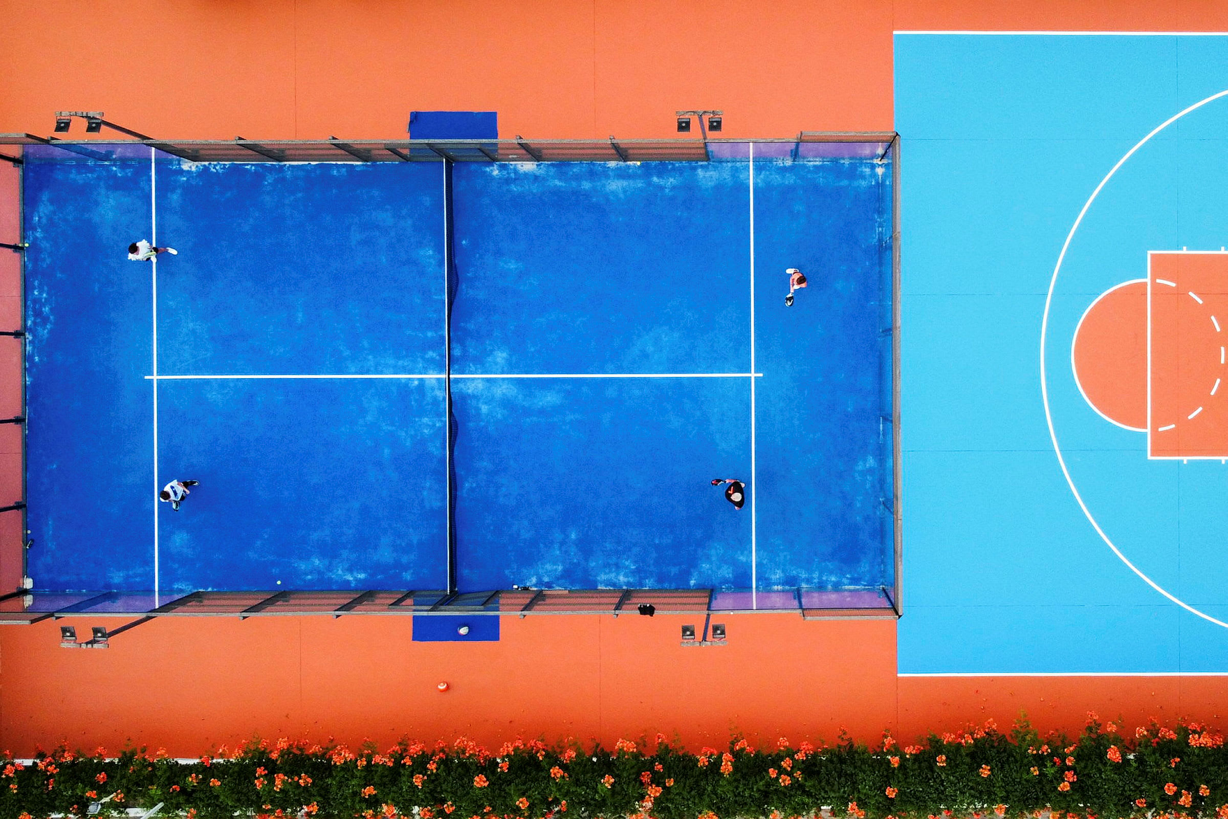 Luftfoto af en blå padelbane med folk, der spiller, ved siden af en orange og blå basketballbane. (AI-genereret tekst)