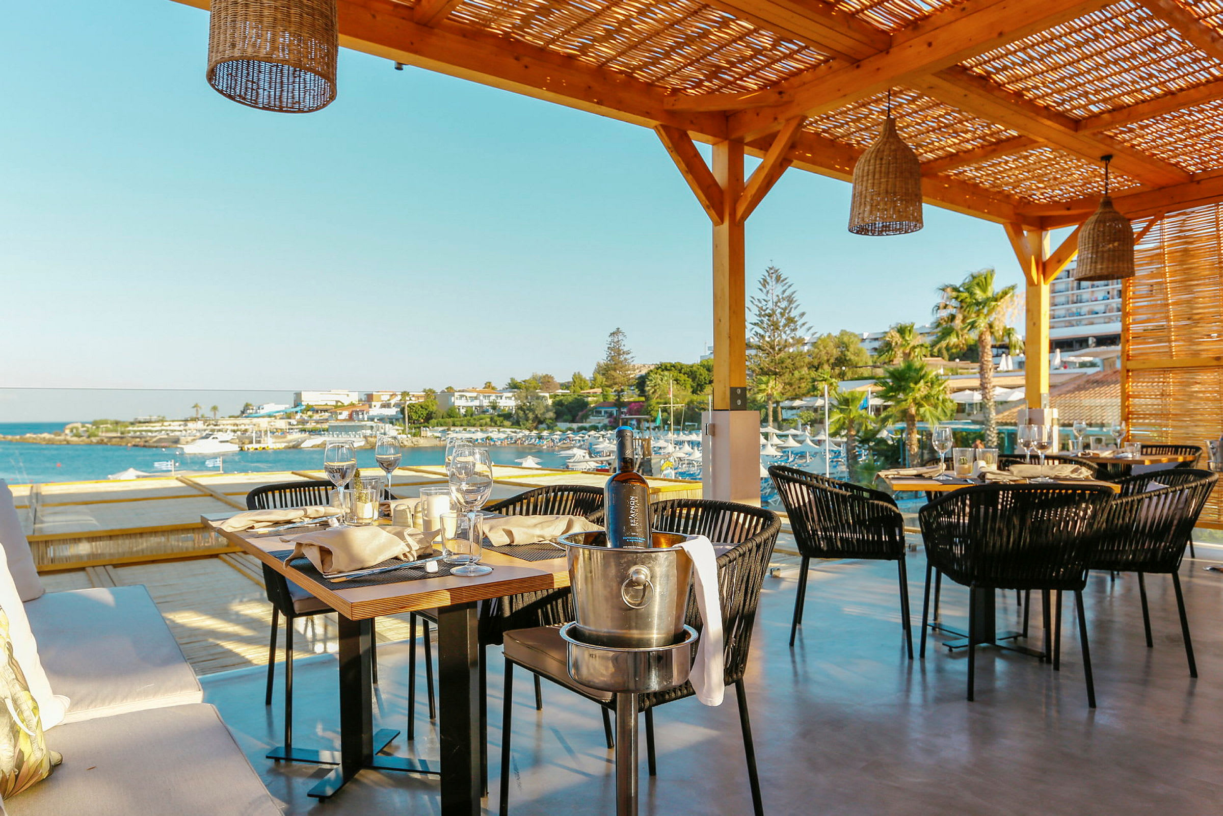 Udendørs serveringsområde med borde og stole under en pergola, med udsigt over havet og stranden. (AI-genereret tekst)