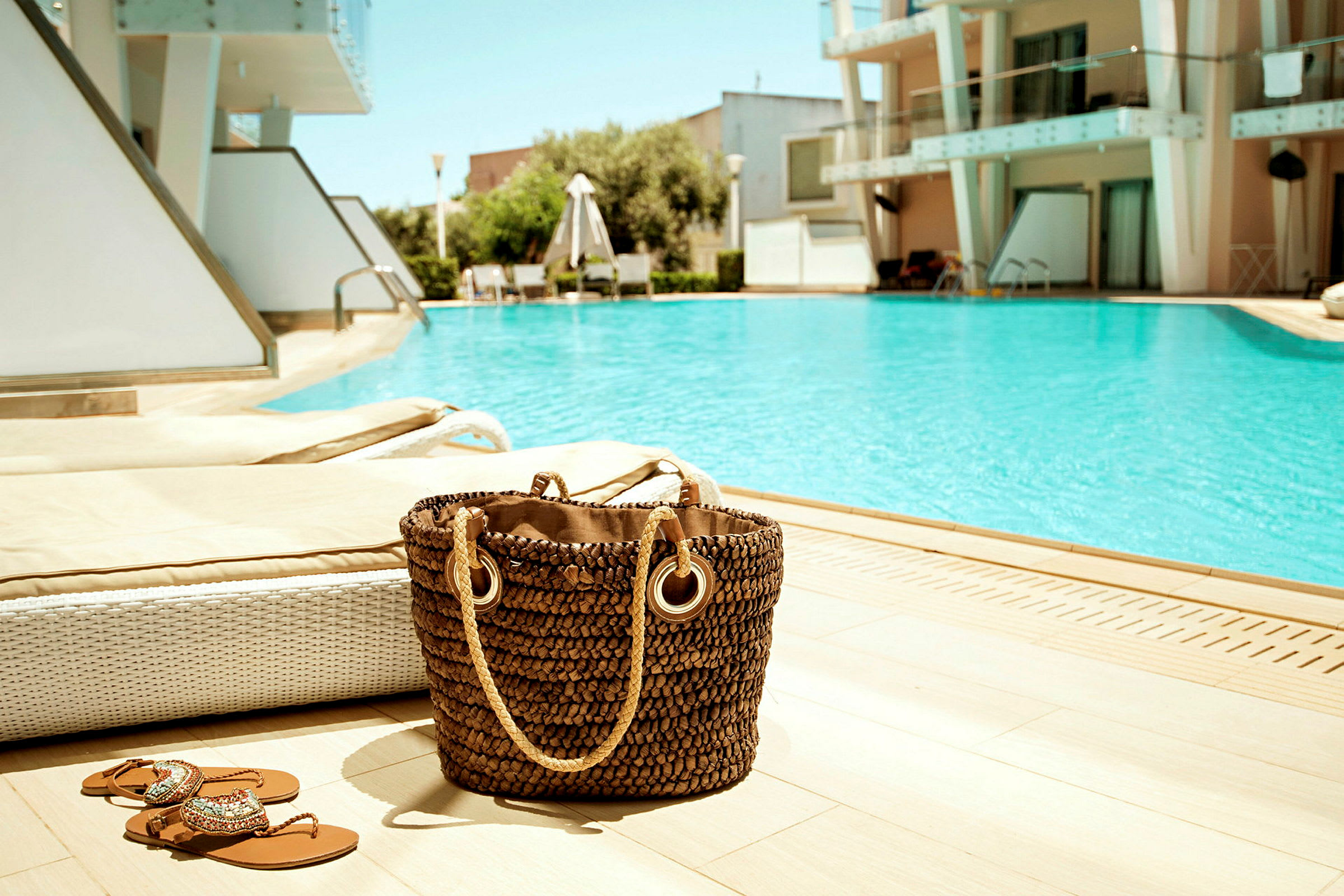 Strandtaske og sandaler ved en swimmingpool, omgivet af solsenge og en moderne bygning i baggrunden. (AI-genereret tekst)