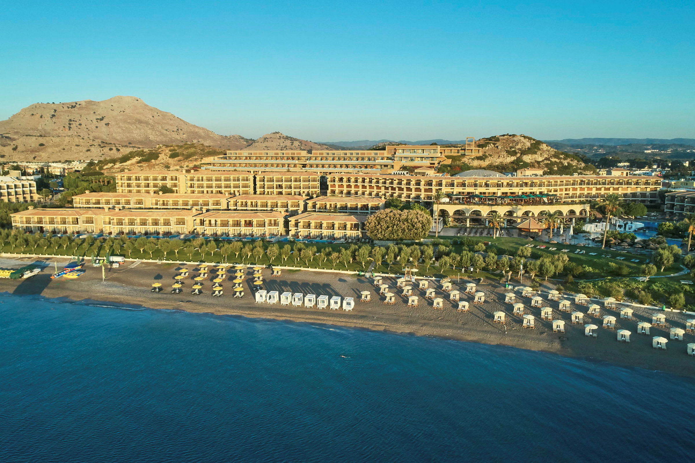 Ved en solrig kyst ligger et stort hotel med flere terrasser og rækker af solsenge og parasoller på stranden. (AI-genereret tekst)