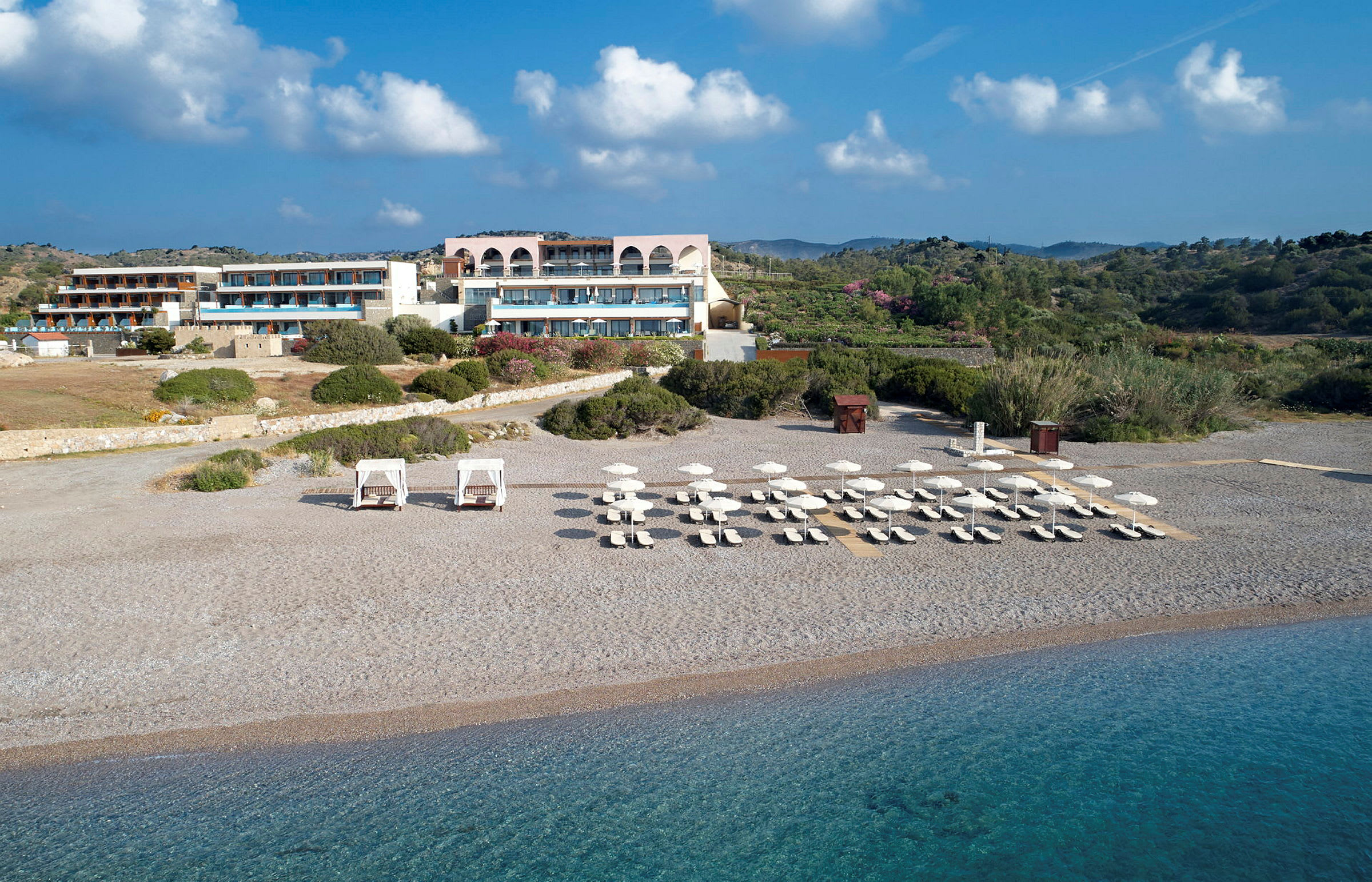 Et strandhotel med liggestole og parasoller opstillede på en stenstrand tæt på klart blåt vand. (AI-genereret tekst)